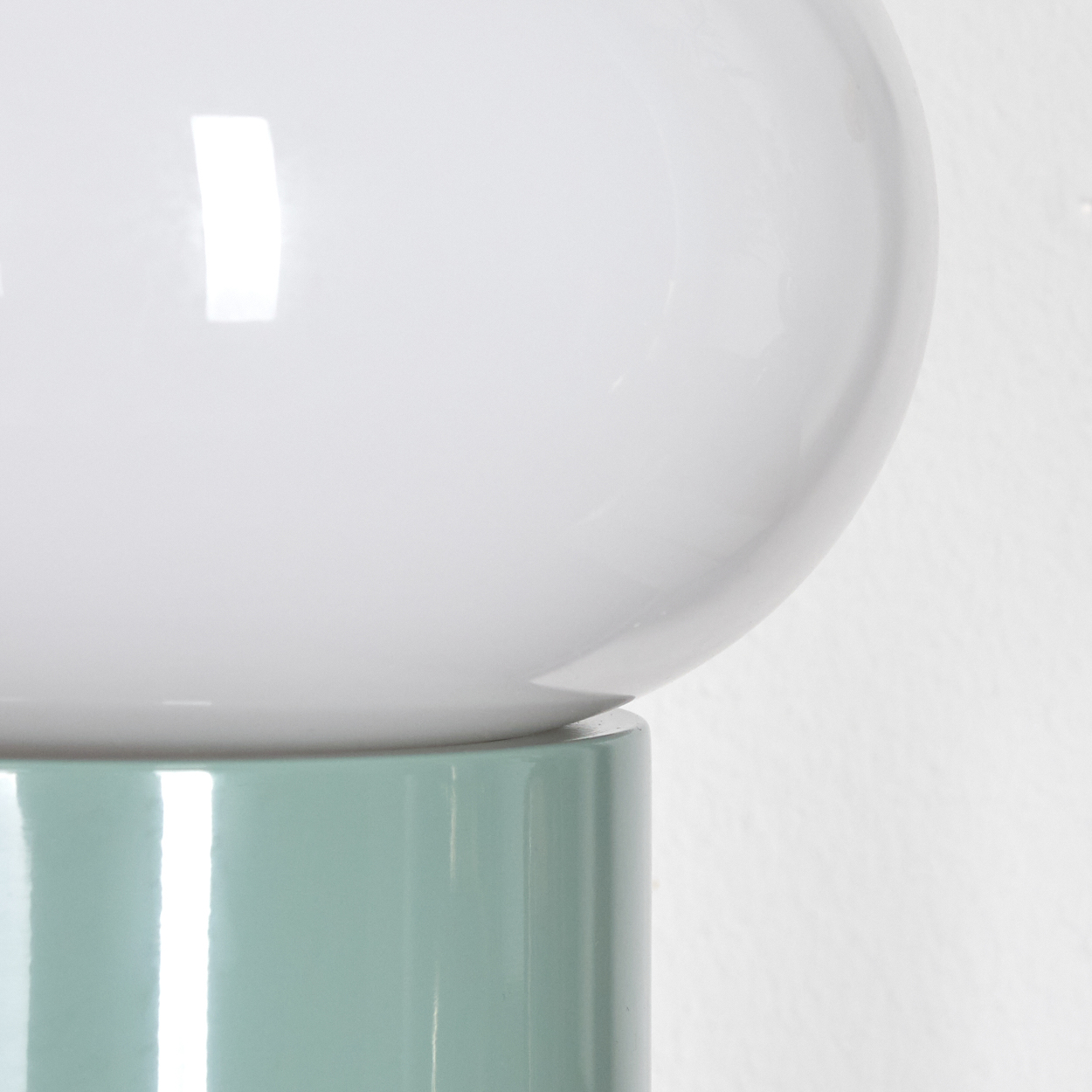 Eine moderne Lampe mit einem weißen, bauchigen Oberteil und einem zylindrischen, hellgrünen Sockel vor einer weißen Wand.