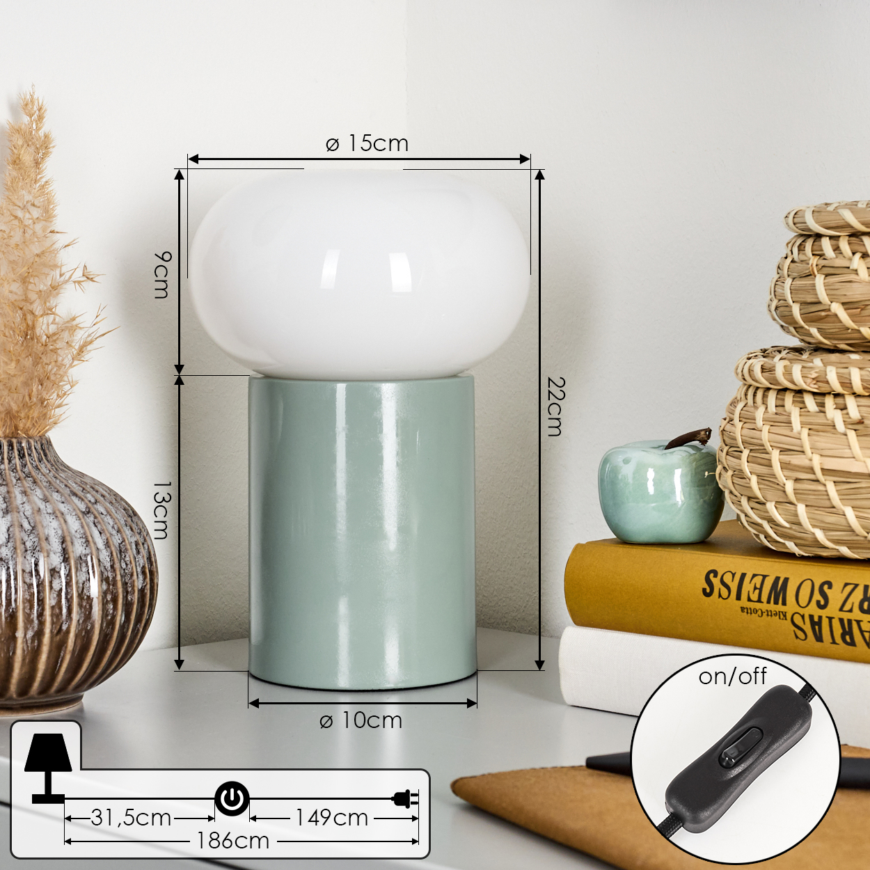 Eine Tischlampe mit weißem Lampenschirm und grünem Sockel. Daneben Bücher, eine Vase, ein kleiner Apfel und ein Korb.
