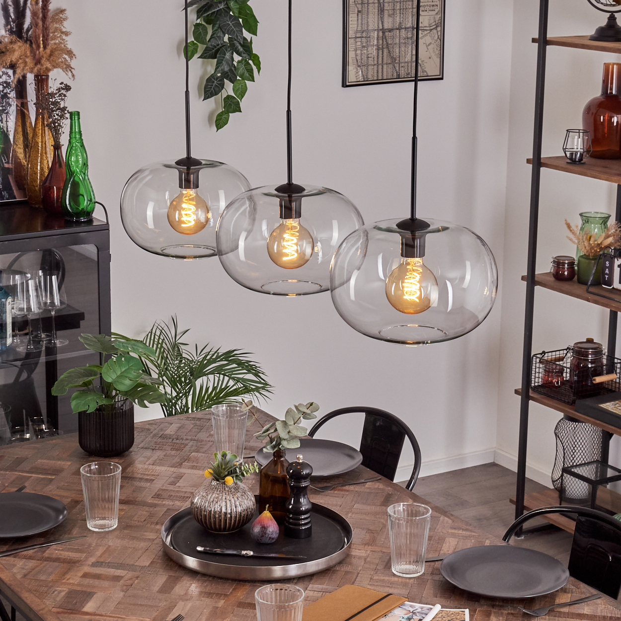 Drei Glaskugel-Pendelleuchten über einem Holztisch, gedeckt. Schwarze Stühle und Regale im Hintergrund.