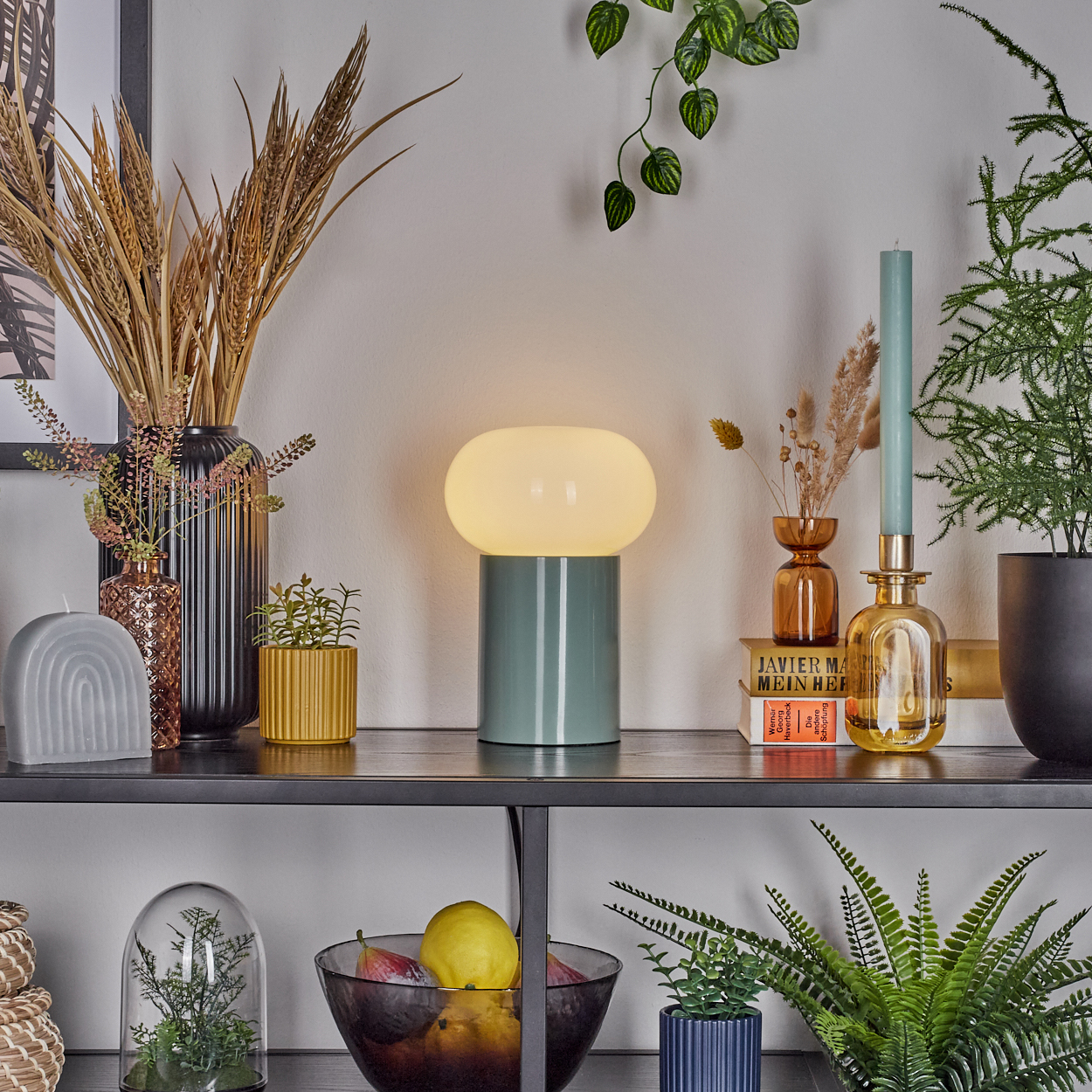Auf einem Regal stehen eine Lampe, Vasen mit Pflanzen, Kerzen und eine Obstschale. Grün-, Gelb- und Brauntöne dominieren.