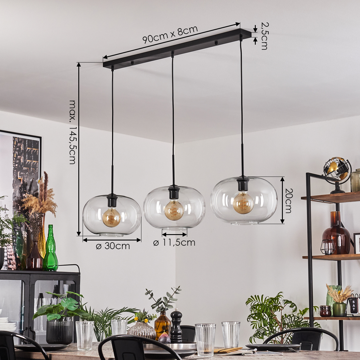 Drei klare Glas-Pendelleuchten hängen über einem Esstisch. Abmessungen sind beschriftet. Der Hintergrund umfasst Deko und ein schwarzes Metallregal.