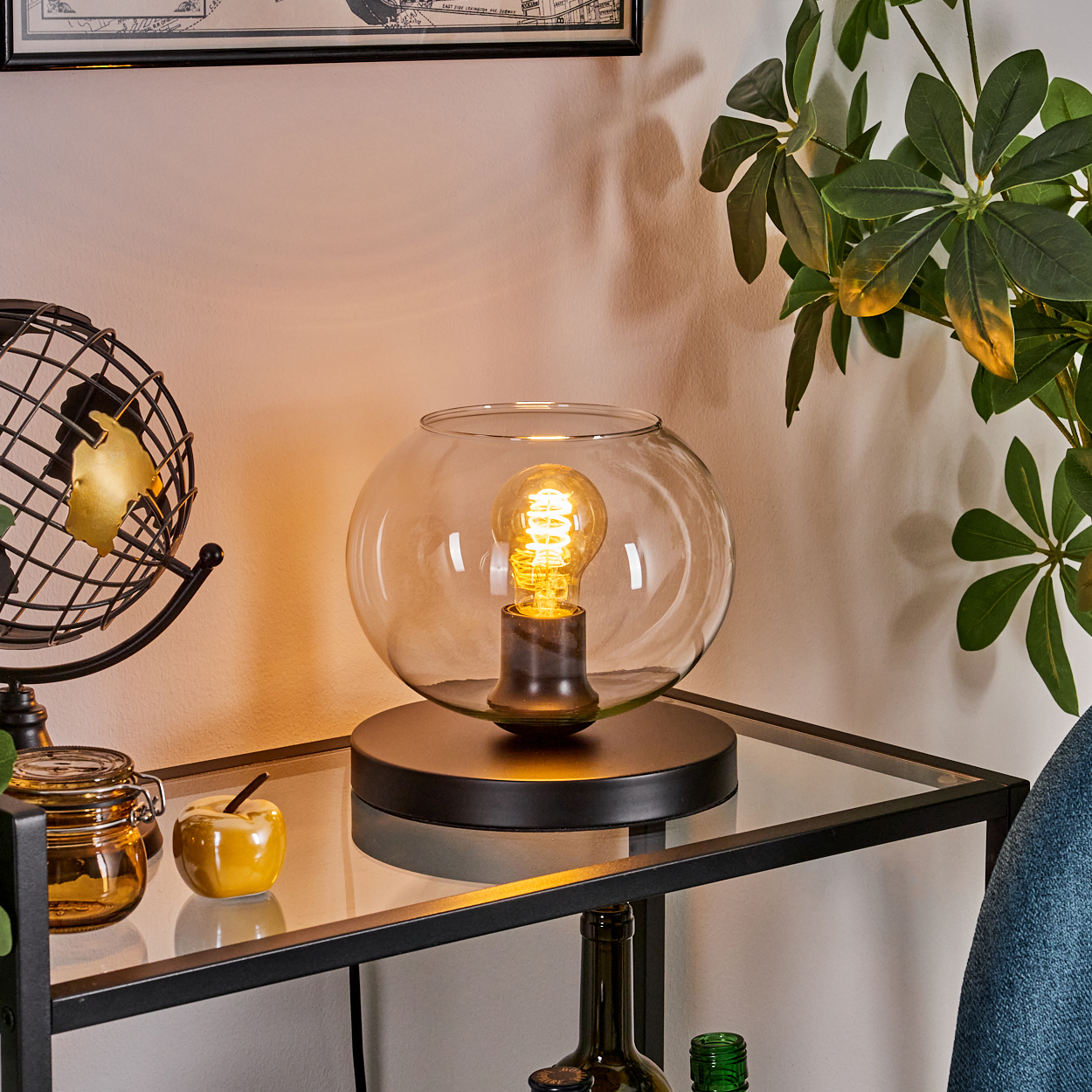 Eine Lampe steht auf einem Glasregal. Globus, Apfel und Pflanze sind in der Nähe. Die Lampe hat einen runden Glasschirm und eine sichtbare Glühbirne.