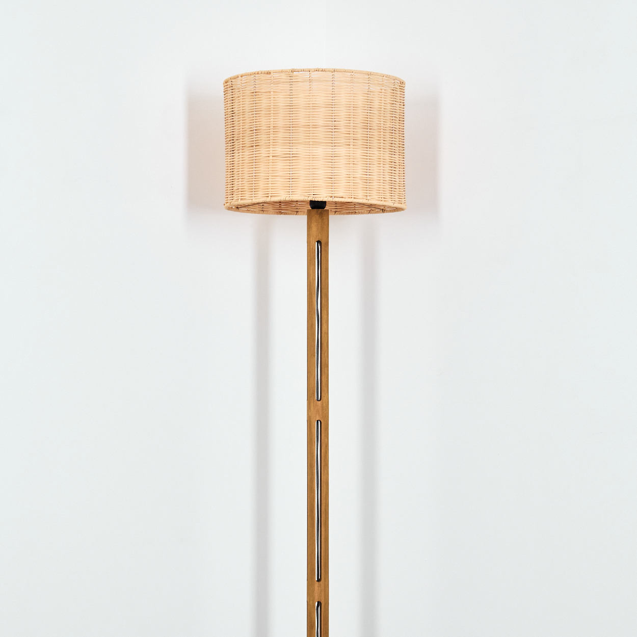 Ein gewebter Lampenschirm sitzt auf einem hölzernen Lampenständer. Die Lampe steht vor einer weißen Wand.