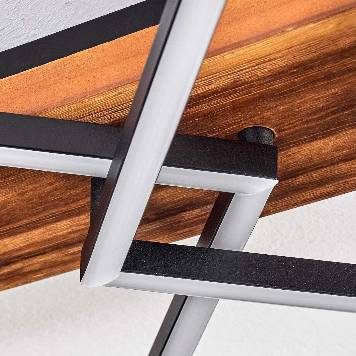 Nahaufnahme der Unterseite eines Tisches. Braune Holzplatte, schwarzer Metallrahmen und weiße Metallstützen kreuzen sich. Weiße Wand im Hintergrund.