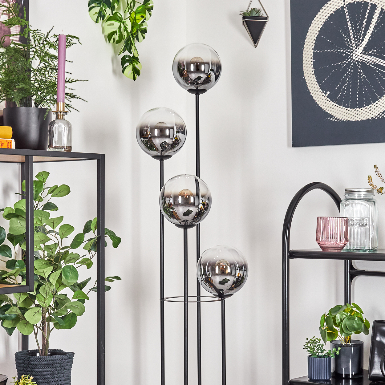 Ein modernes Wohnzimmer mit einer Stehlampe mit vier runden Glasschirmen, Pflanzen und Dekorationsartikeln. Die Wände sind weiß.