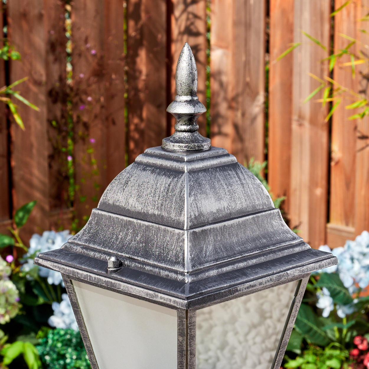 Eine dekorative Außenlampe mit silbernem Oberteil, vor einem Holzzaun und Grün. Die Lampe hat eine pyramidenförmige Spitze.