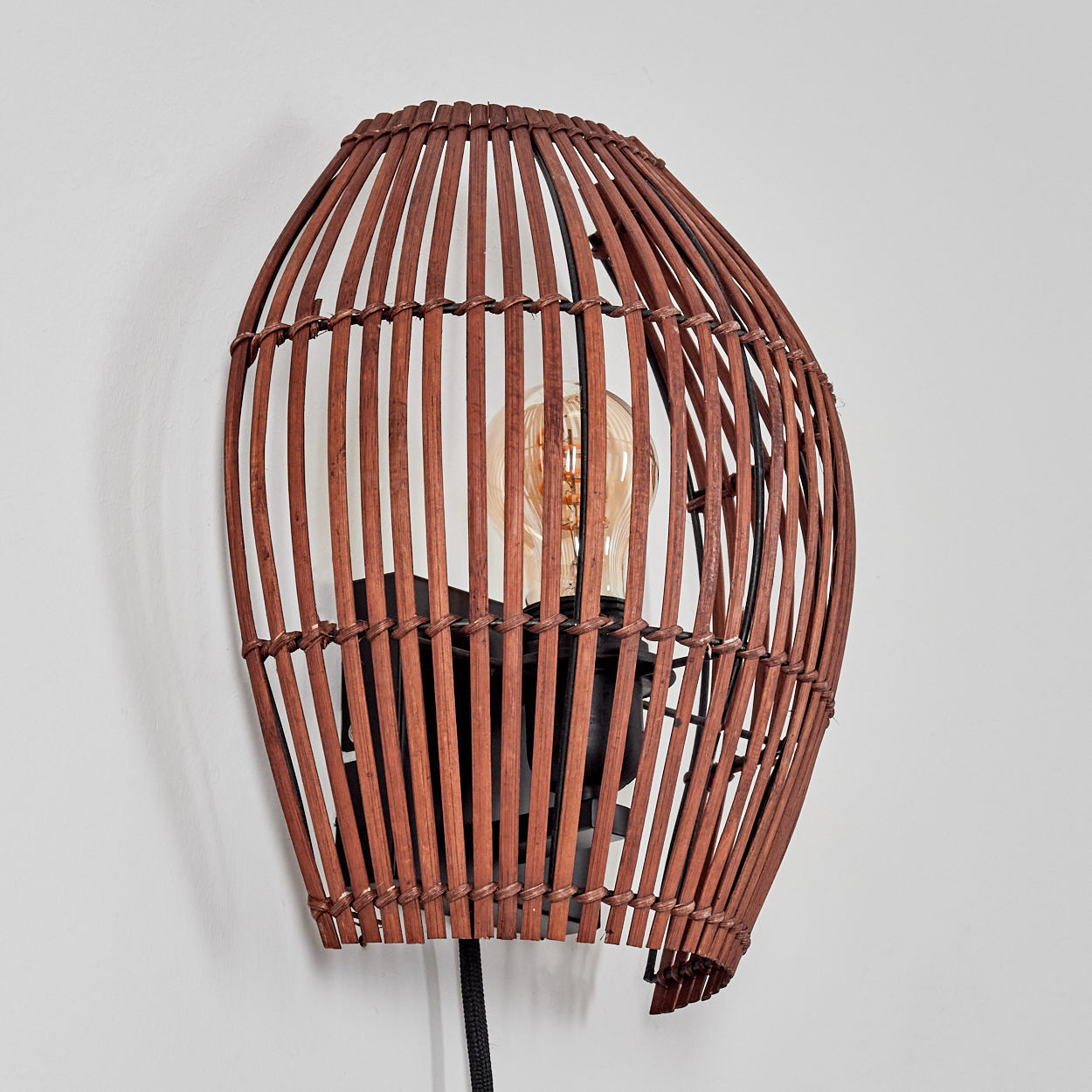 Eine braune Korblampe ist an einer weißen Wand montiert. Die Lampe hat eine sichtbare Glühbirne. Ein schwarzes Kabel hängt herunter.