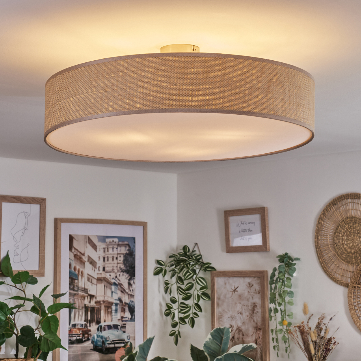 Eine große, runde Deckenlampe mit braunem Stoffschirm hängt über gerahmten Bildern und Pflanzen. Der Raum ist gut beleuchtet und dekoriert.