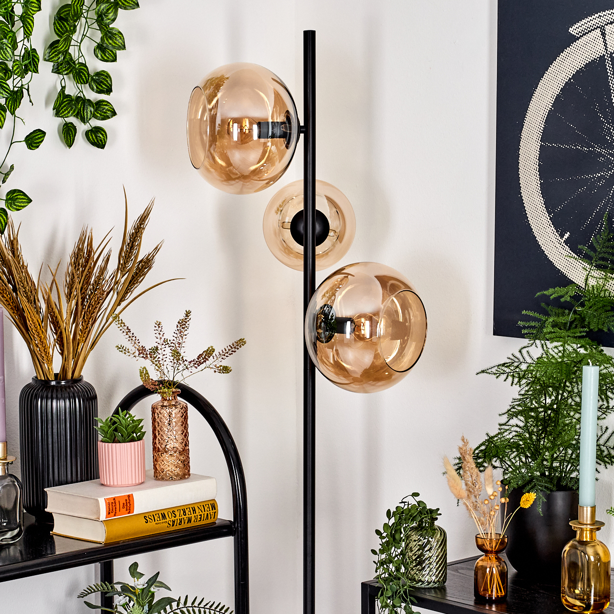 Eine moderne Stehlampe mit Glaskugeln steht neben Pflanzen und Dekoration. Ein Fahrradrad-Kunstwerk ist an einer weißen Wand zu sehen.