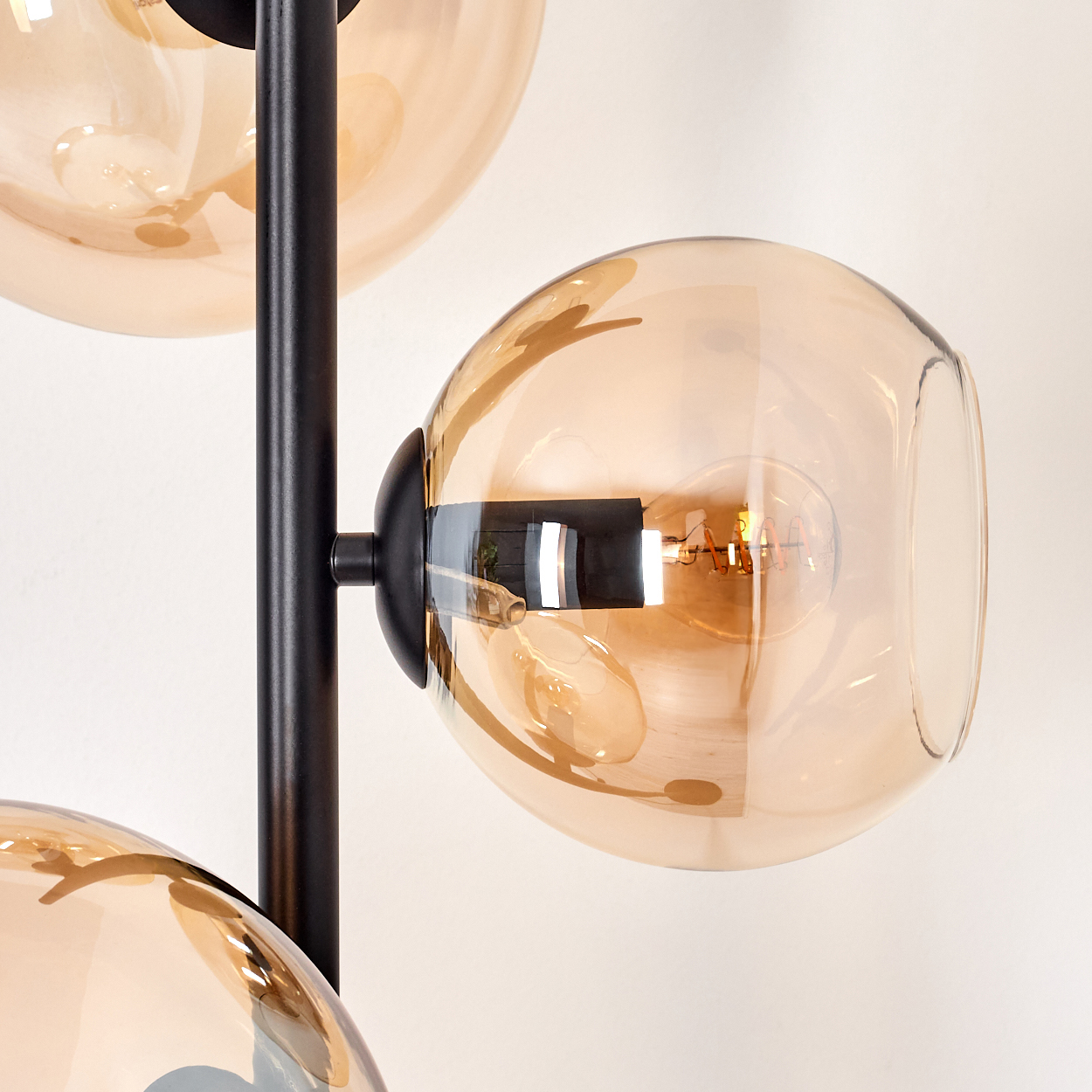Nahaufnahme einer modernen Lampe mit drei bernsteinfarbenen Glaskugeln. Schwarzer Metallrahmen. Glühbirnen sind in den Kugeln sichtbar. Weißer Hintergrund.