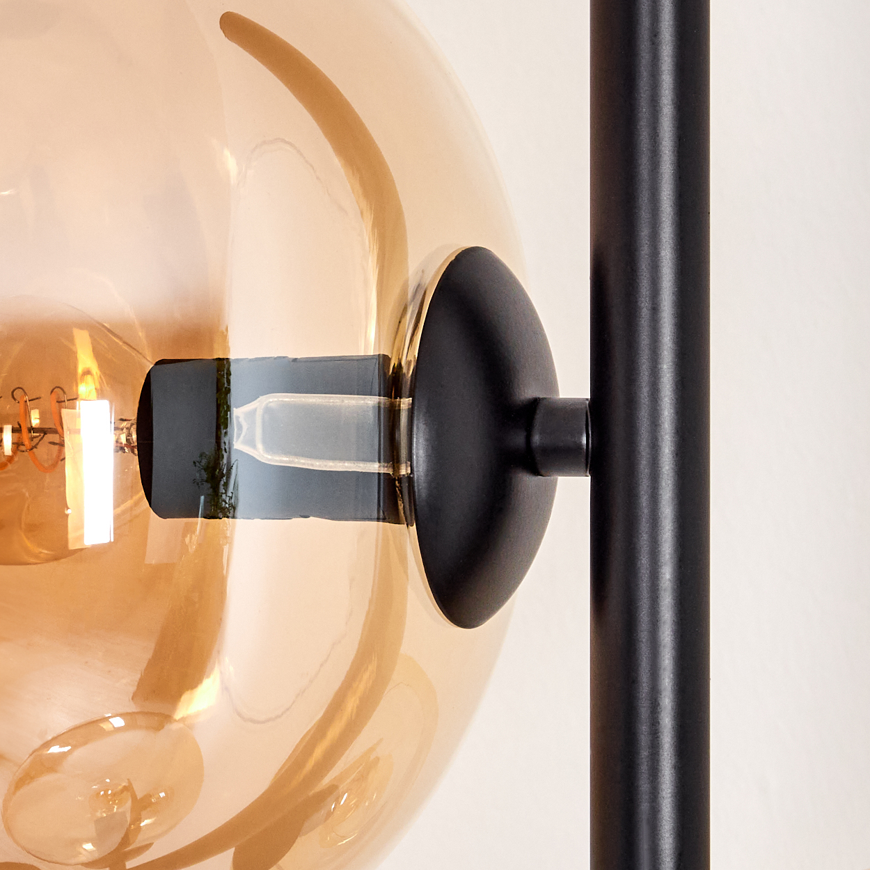 Nahaufnahme einer Lampe mit bernsteinfarbenem, kugelförmigem Glasschirm, schwarzer Halterung und schwarzer Stütze vor weißem Hintergrund.
