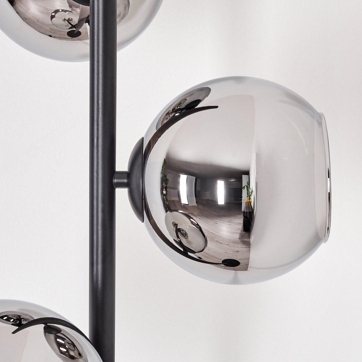 Eine moderne Lampe mit drei reflektierenden, kugelförmigen Schirmen. Ein schwarzer Pfosten trägt sie vor einer weißen Wand. Die Schirme spiegeln einen Raum wider.