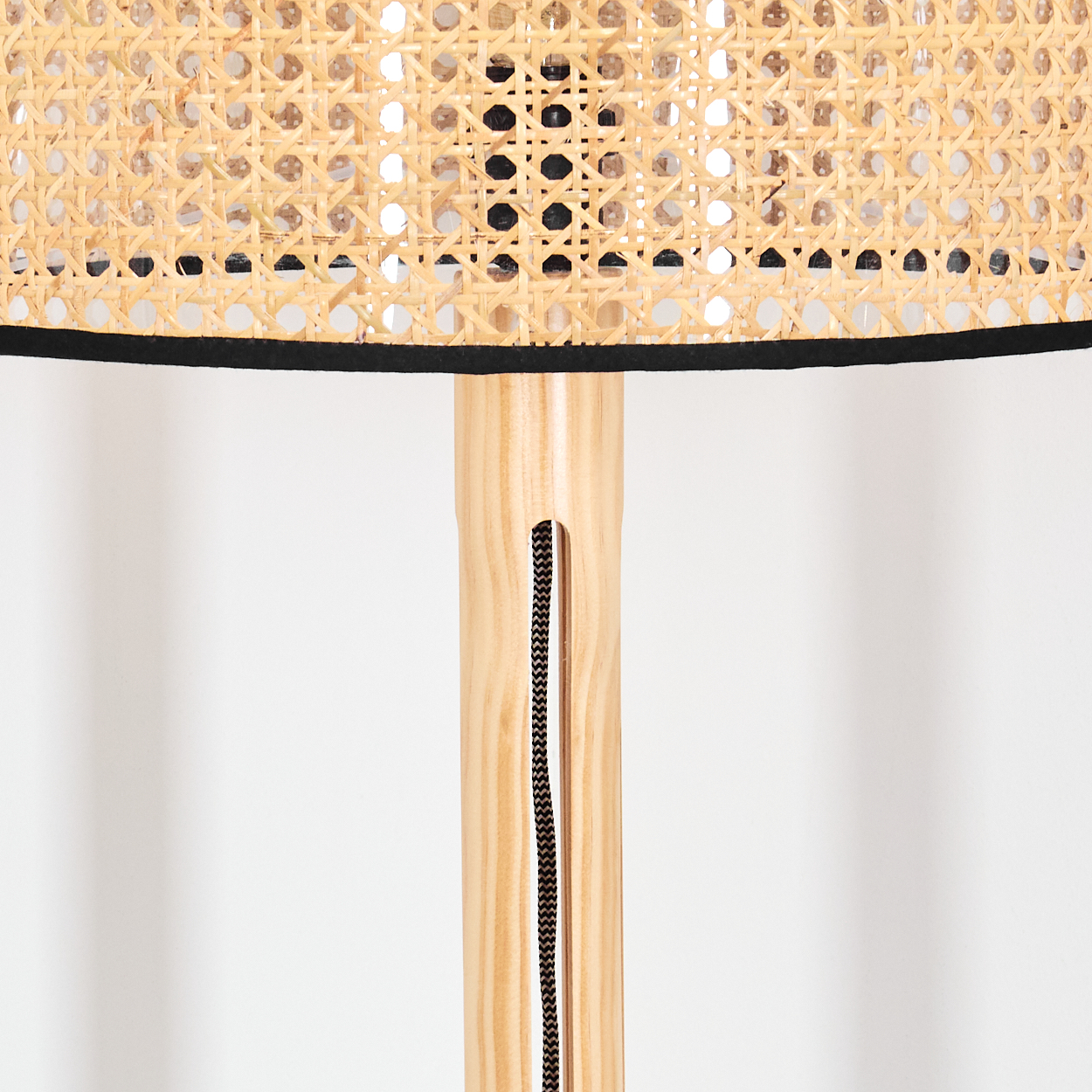 Nahaufnahme einer Lampe. Lampenschirm aus gewebtem Rattan. Schwarze Zierleiste. Heller Holzfuß. Schwarzes Kabel verläuft durch den Fuß.