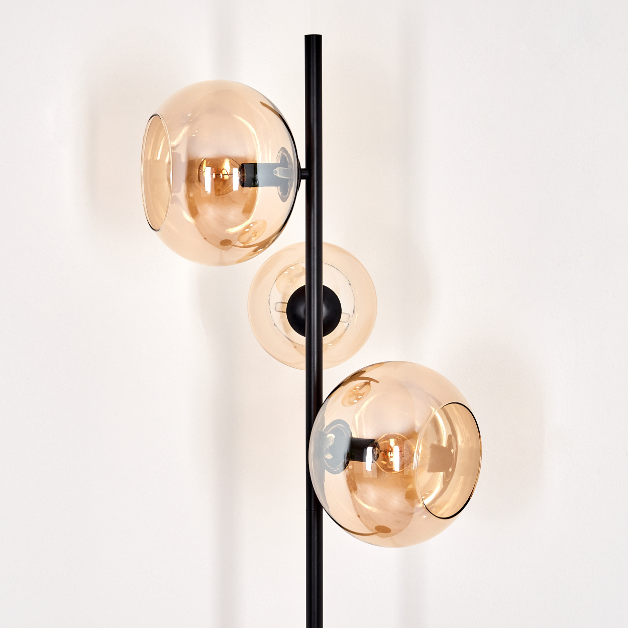 Eine moderne Stehlampe mit drei Glaskugelschirmen in Bernstein, vor einer weißen Wand. Die Lampe hat einen schwarzen Metallrahmen.