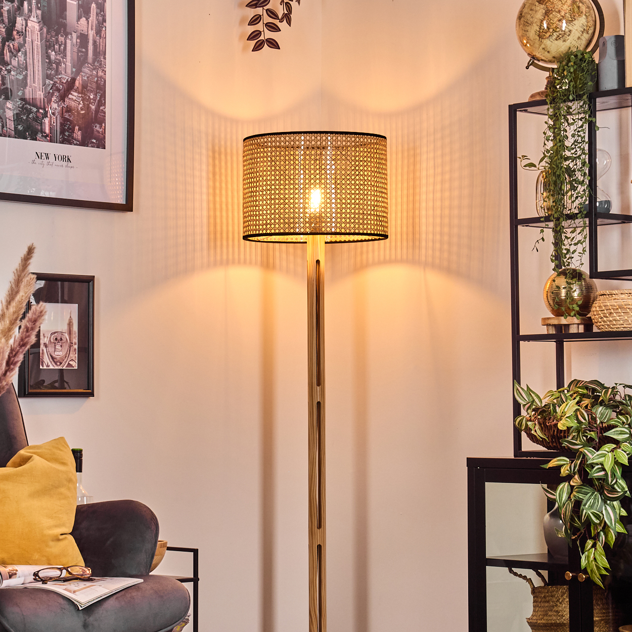 Eine beleuchtete Stehlampe aus Holz steht in einem Raum. Im Hintergrund sind ein gerahmtes New York-Bild und ein Regal mit Pflanzen.