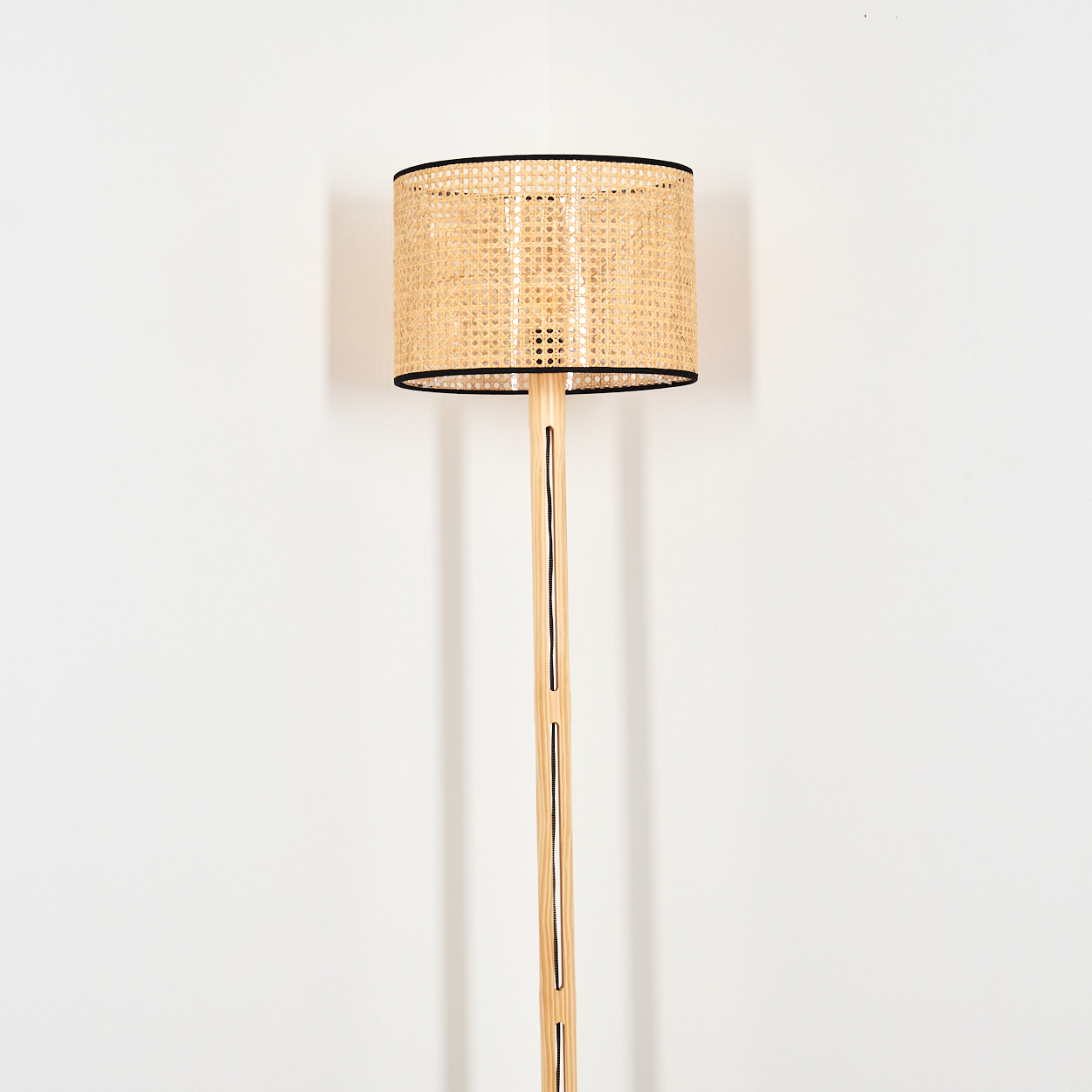Eine Stehlampe steht vor einer weißen Wand. Sie hat einen gewebten Lampenschirm mit schwarzem Rand und eine helle Holzstange.