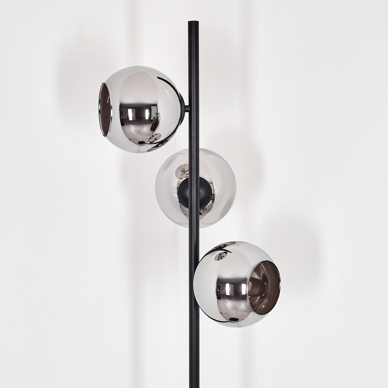 Eine moderne Stehlampe mit drei kugelförmigen Schirmen. Zwei sind verchromt, einer ist klar. Das Gestell ist schwarz, vor einer weißen Wand.