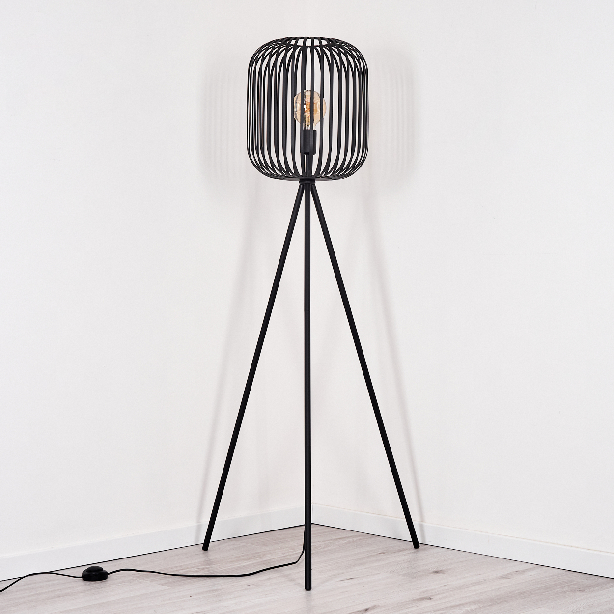 Eine schwarze Metall-Stativ-Stehlampe mit käfigartigem Lampenschirm. Die Glühbirne ist sichtbar. Steht in einem weißen Raum.