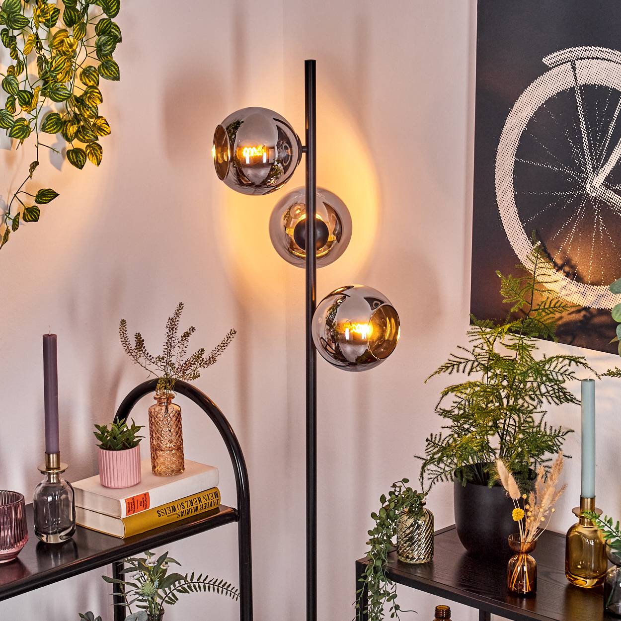 Ein modernes Zimmer mit einer Stehlampe mit drei Kugelschirmen, Pflanzen, Regalen mit Dekor und einem Fahrradrad-Kunstwerk.