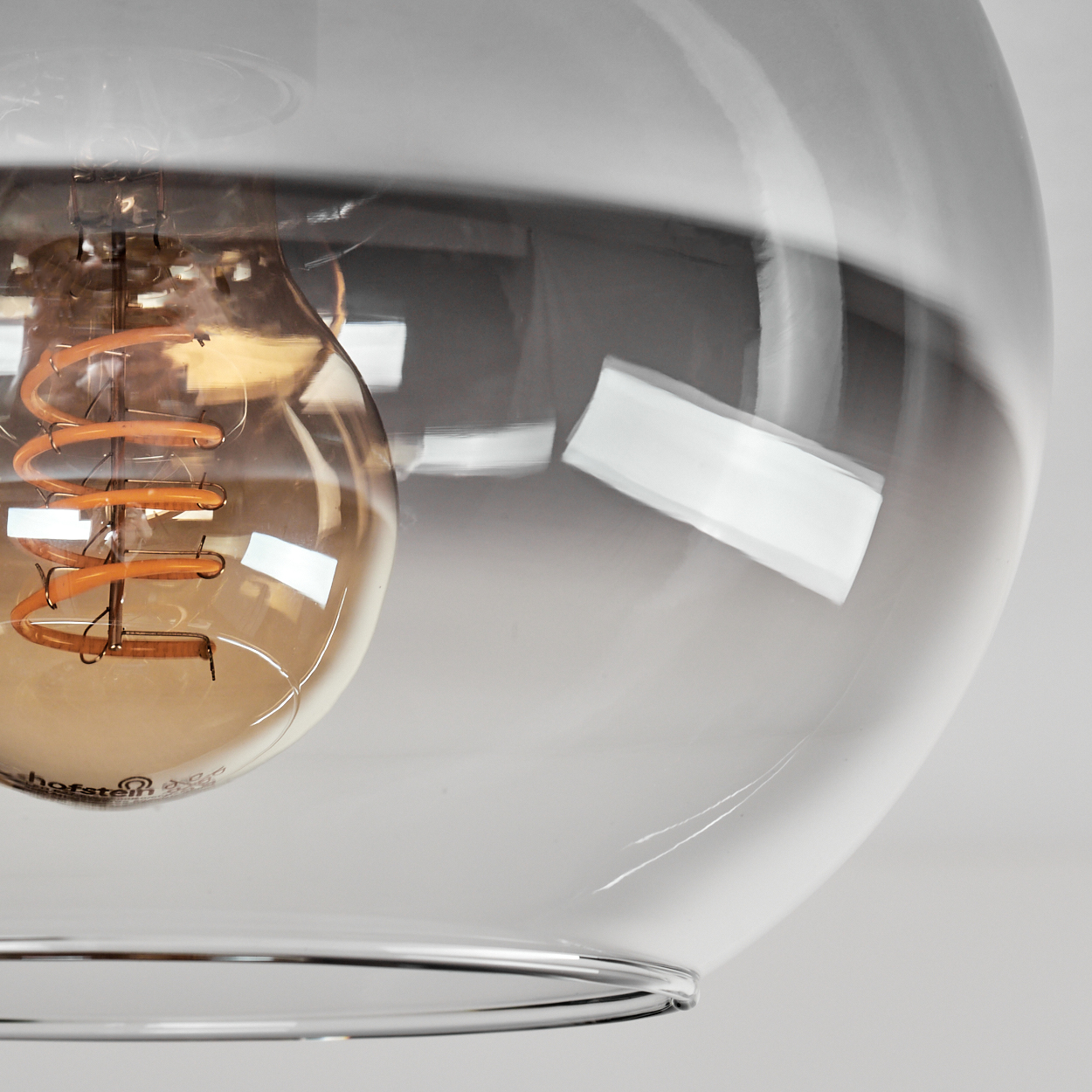 Nahaufnahme einer Glas-Pendelleuchte. Eine warmtonige Glühbirne befindet sich in einer transparenten Glaskugel. Der Glühfaden ist sichtbar.