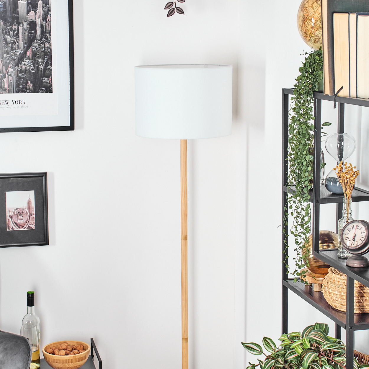 Eine weiße Lampe steht vor einer weißen Wand. Rechts ein Regal voller Dekorationen und Pflanzen. Links Bilder und eine Holzschale.