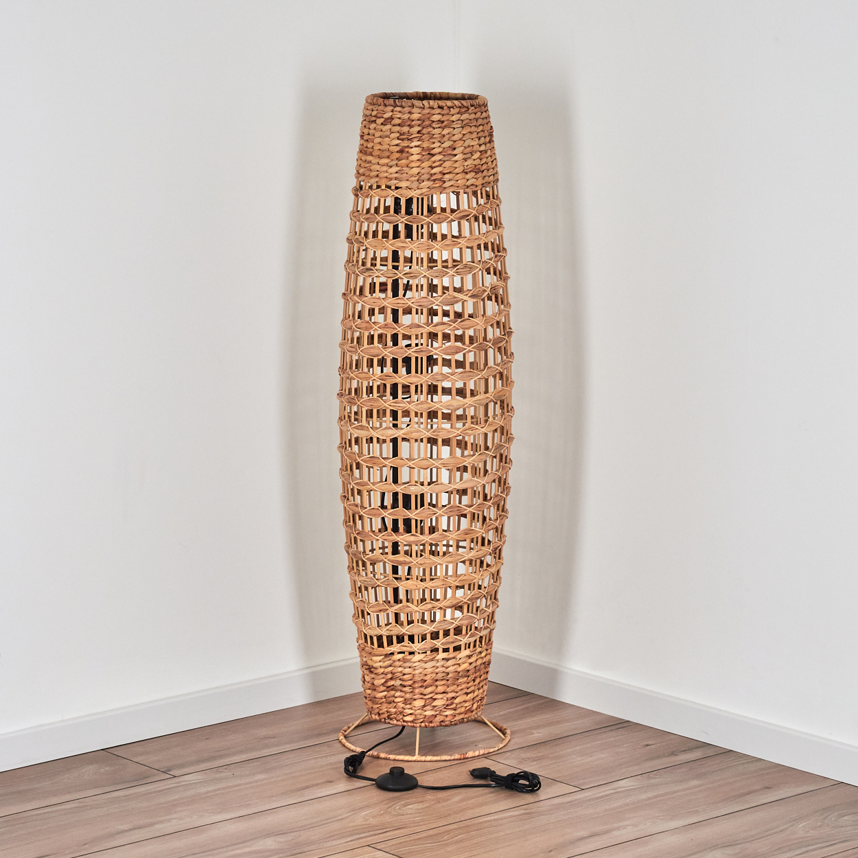 Eine hohe Stehlampe aus Korbgeflecht steht in einer Ecke. Die Lampe ist beige und zylindrisch, mit einem Webmuster. Sie steht auf einem Holzboden.