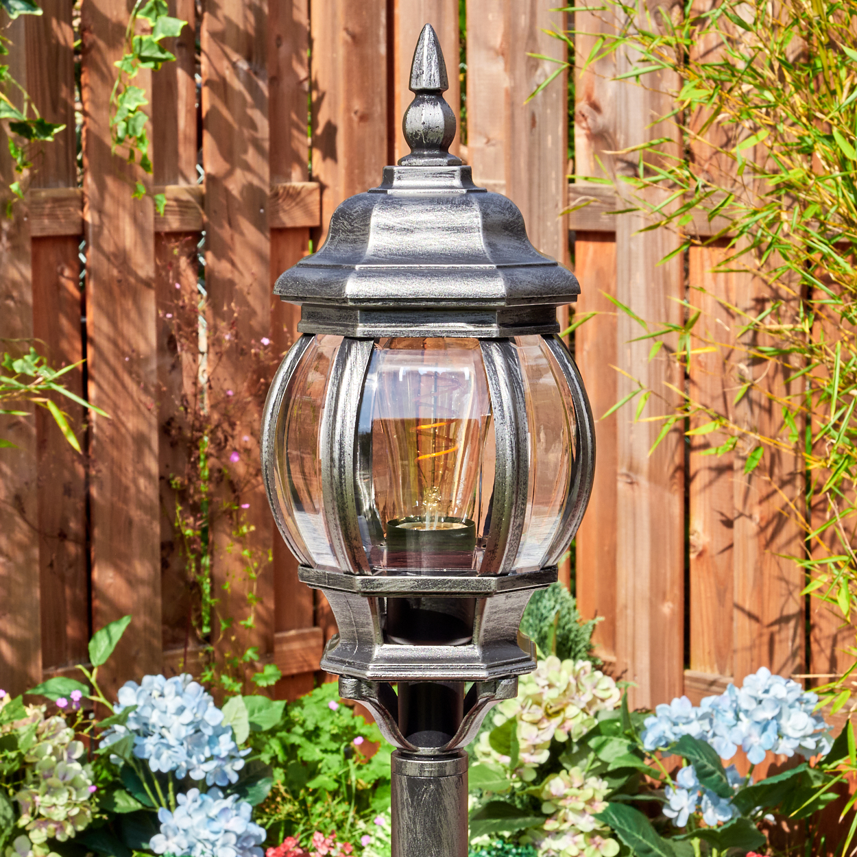 Eine dekorative Außenlampe mit Glaskugel und silbernem Metallrahmen, vor einem Holzzaun und Blumen.