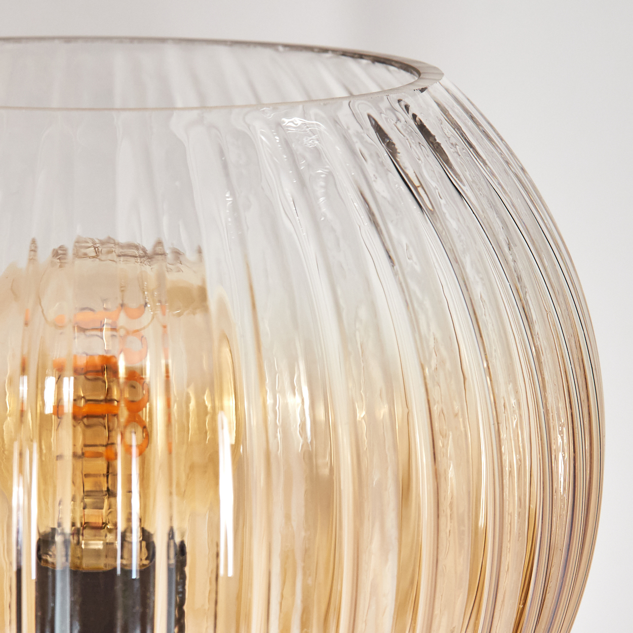 Nahaufnahme eines Lampenschirms aus Glas mit vertikalen Rippen. Im Inneren ist eine Edison-Glühbirne zu sehen. Der Hintergrund ist weiß.