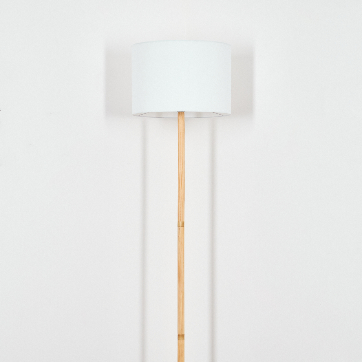 Eine Stehlampe mit weißem Lampenschirm und Holzstiel steht vor einer weißen Wand. Die Lampe wirft einen Schatten.