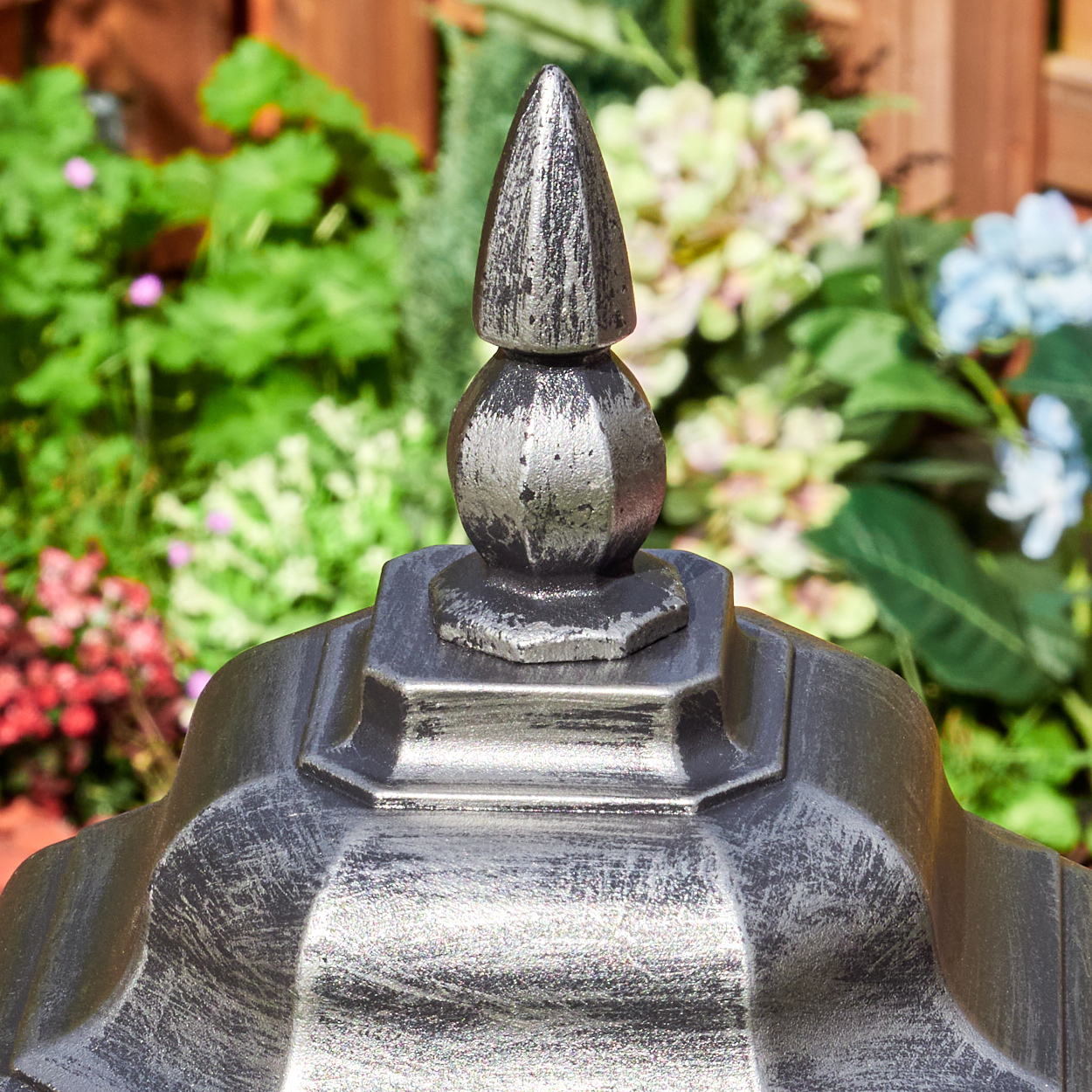 Ein dekoratives Metallobjekt mit spitzem Aufsatz vor einem verschwommenen grünen und floralen Hintergrund.