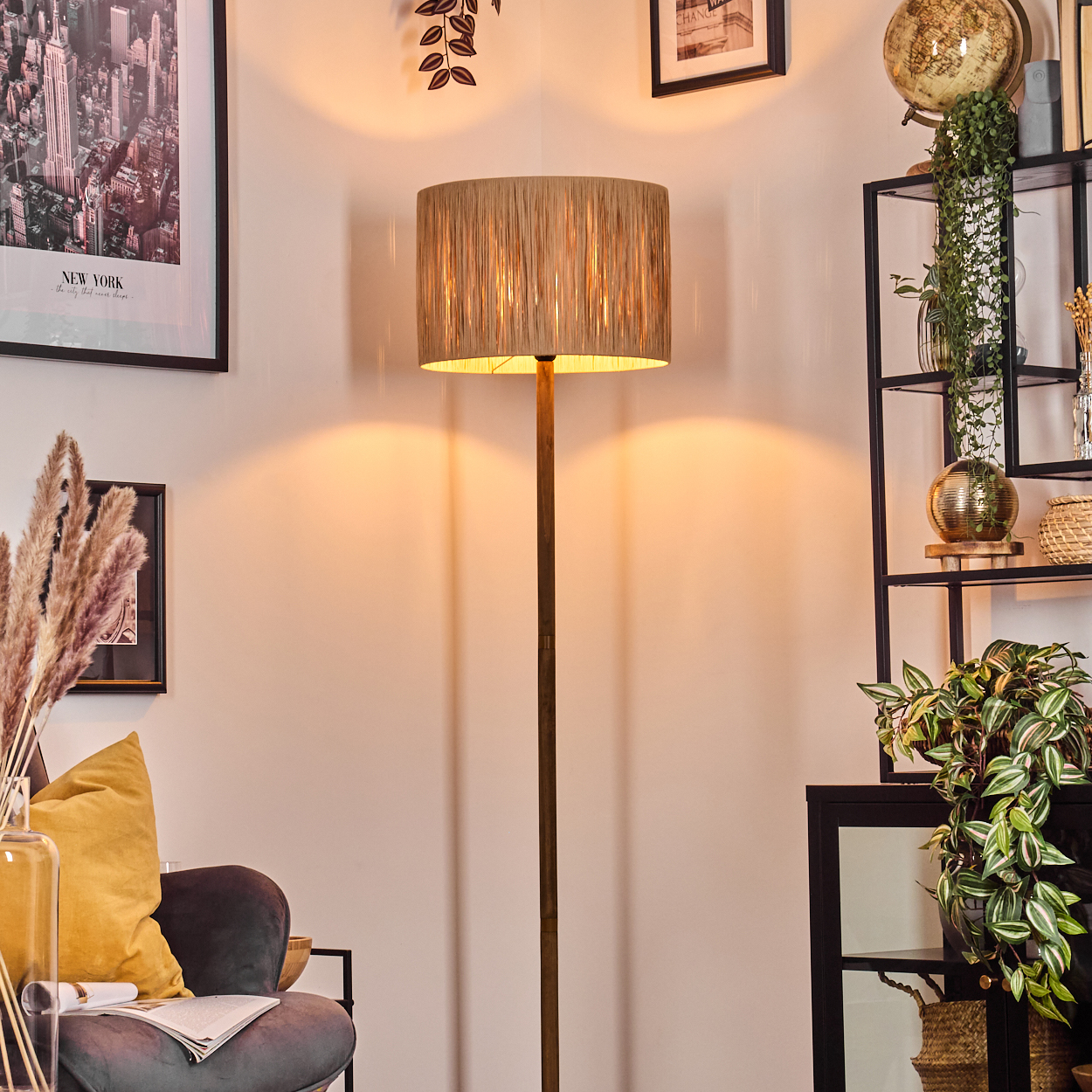 Eine beleuchtete Stehlampe steht in einem Raum mit einem Regal, einem Stuhl und Bildern. Die Lampe hat einen beigen Lampenschirm.
