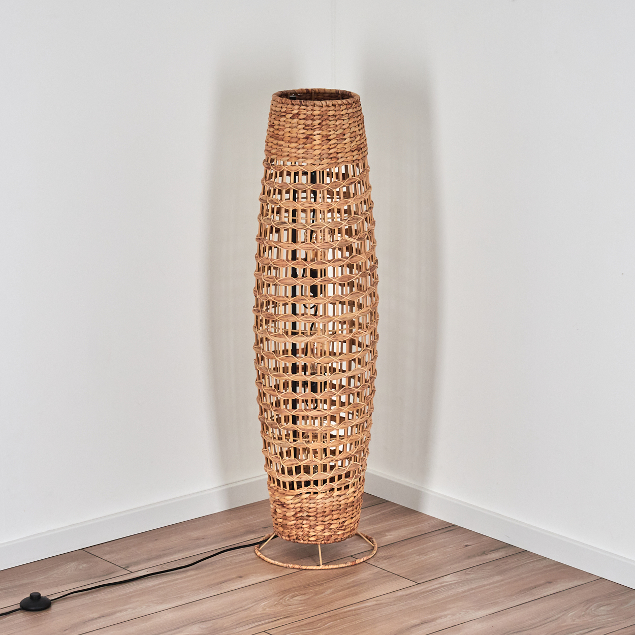 Eine gewebte Stehlampe aus Rattan steht in einer Ecke. Die Lampe ist beige mit einem dunklen Kabel und einem schwarzen Schalter.