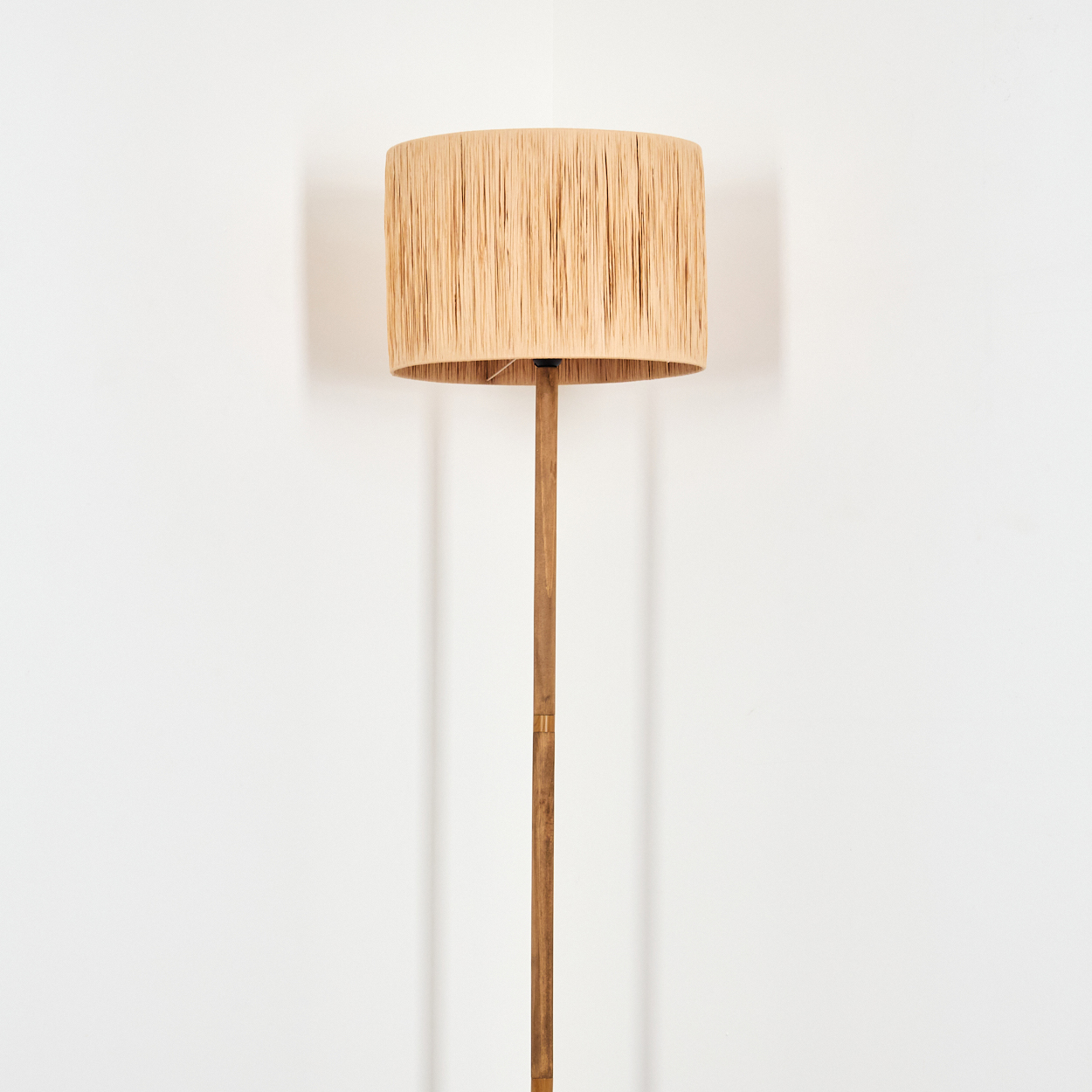 Eine Stehlampe mit beigem Lampenschirm und Holzstiel steht in einer Ecke vor einer weißen Wand.
