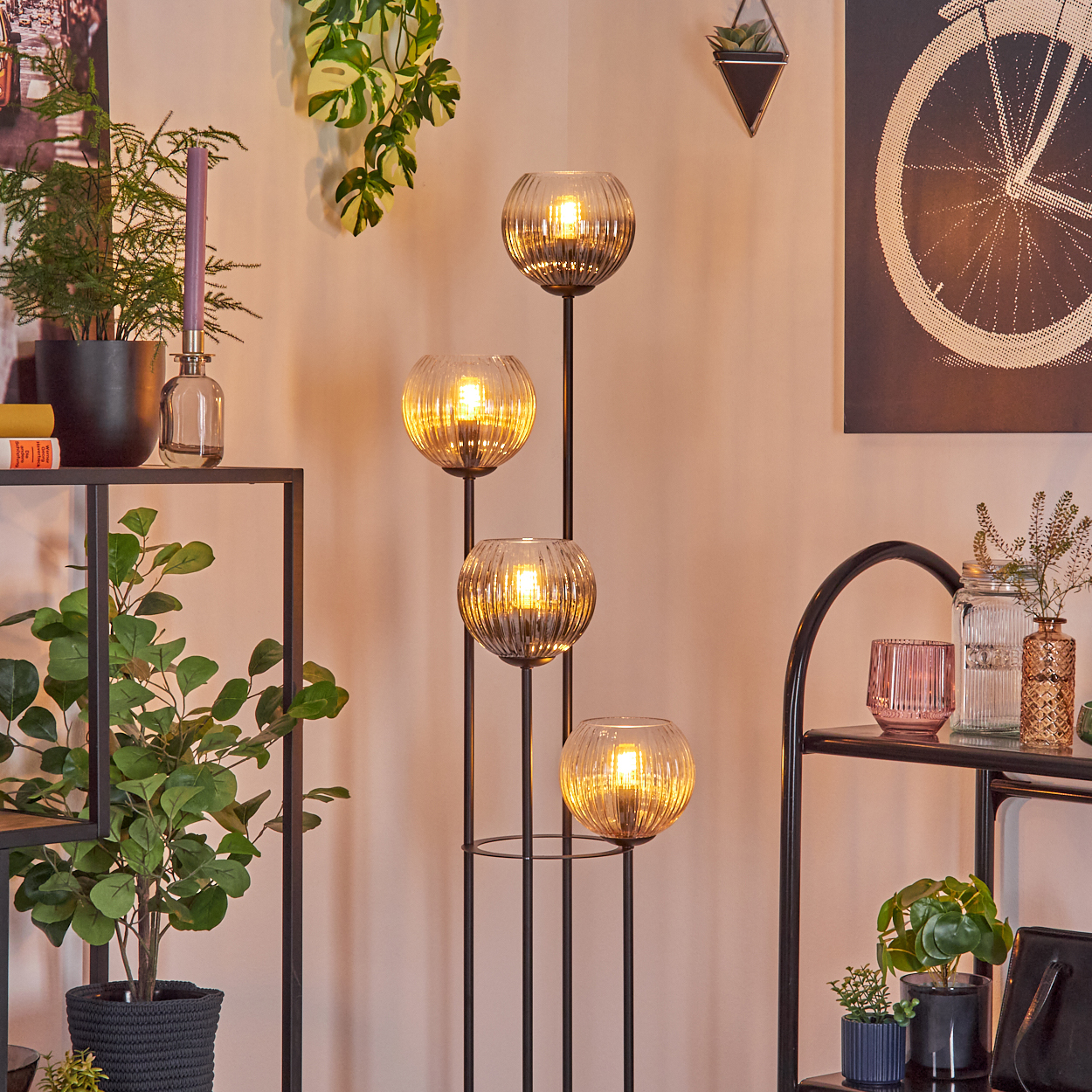 Eine dekorative Lampe mit vier Glaskugeln leuchtet. Pflanzen und Regale mit Dekoration befinden sich im Hintergrund.