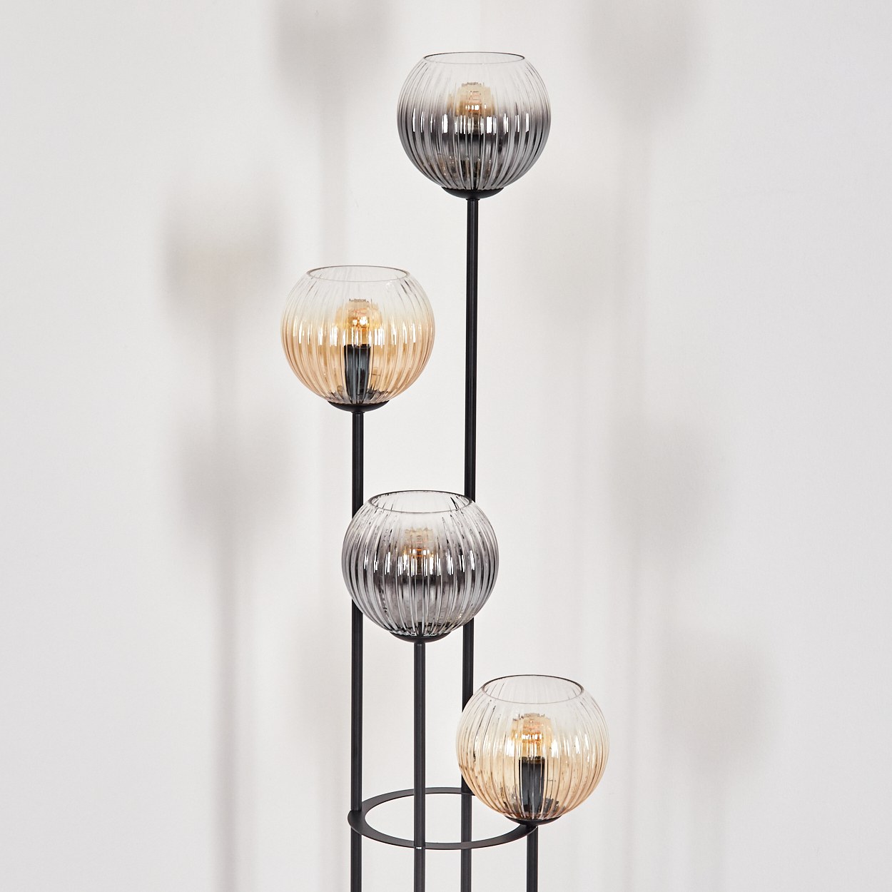 Eine moderne Stehlampe mit vier kugelförmigen Lampenschirmen. Zwei sind bernsteinfarben, zwei grau, auf einem schwarzen Metallrahmen, vor einer weißen Wand.