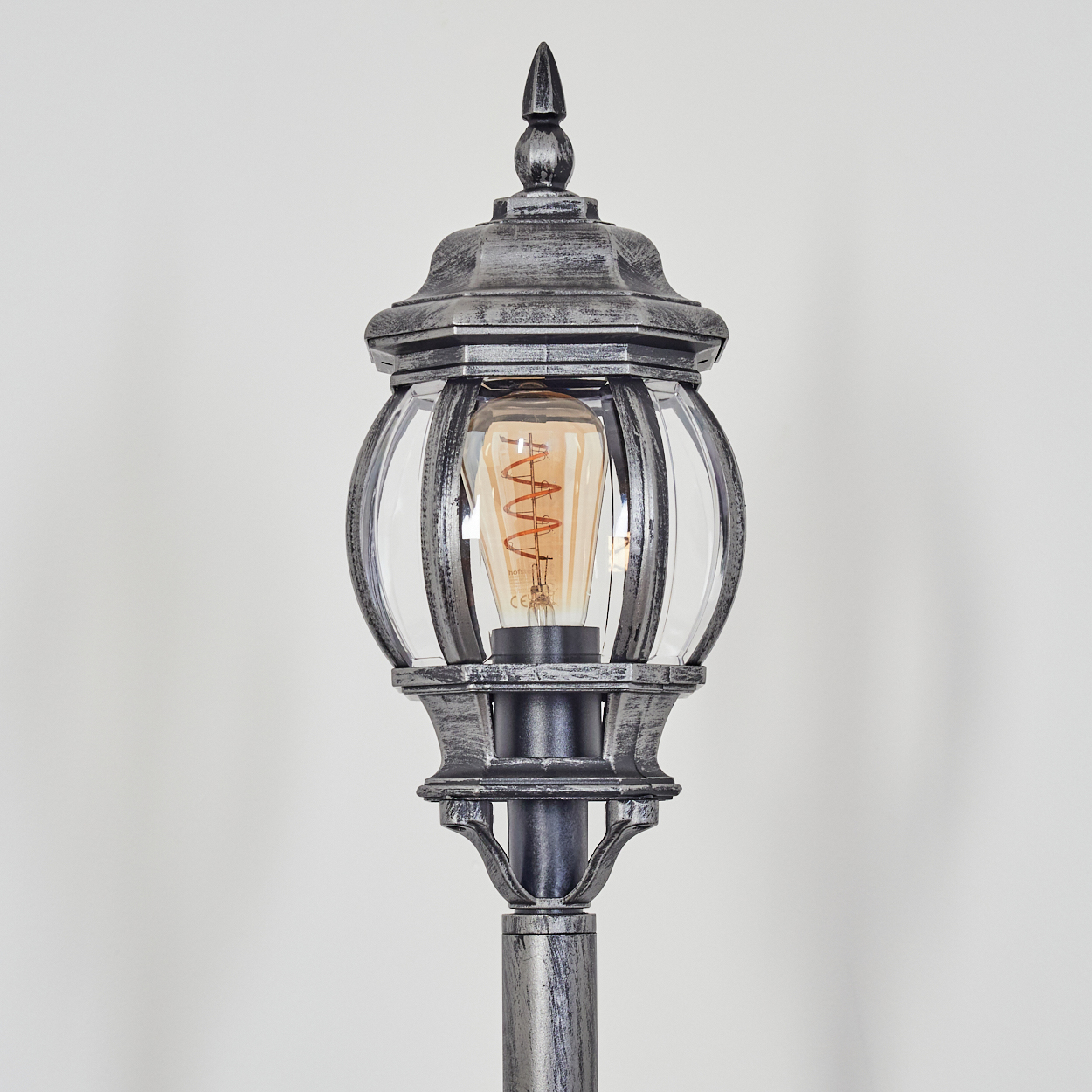 Eine dekorative Außenlaterne. Sie hat eine Glaskugel mit einer Wendelbirne. Der Metallrahmen ist grau und schwarz.