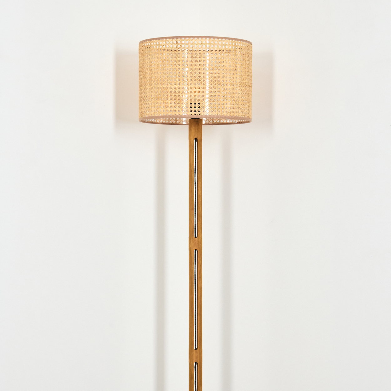 Eine hohe Stehlampe mit gewebtem Lampenschirm und Holzfuß steht vor einer weißen Wand. Der Lampenschirm ist zylindrisch.