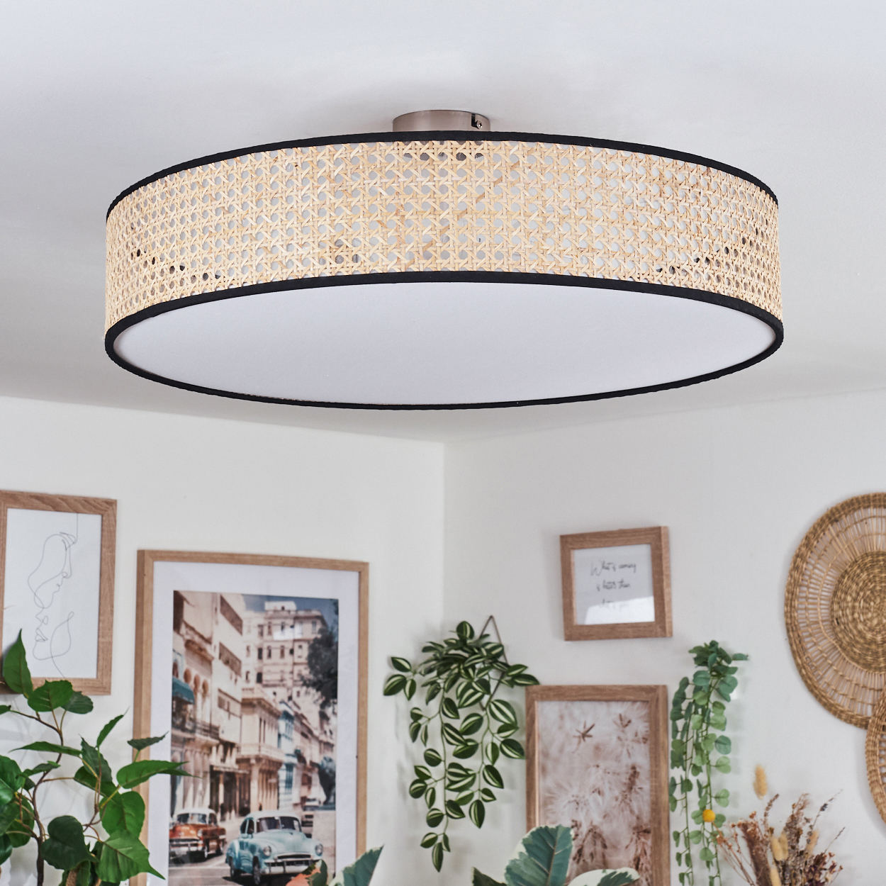 Eine ovale Deckenleuchte mit geflochtenem Rattanschatten. Darunter gerahmte Bilder und Pflanzen vor einer weißen Wand.