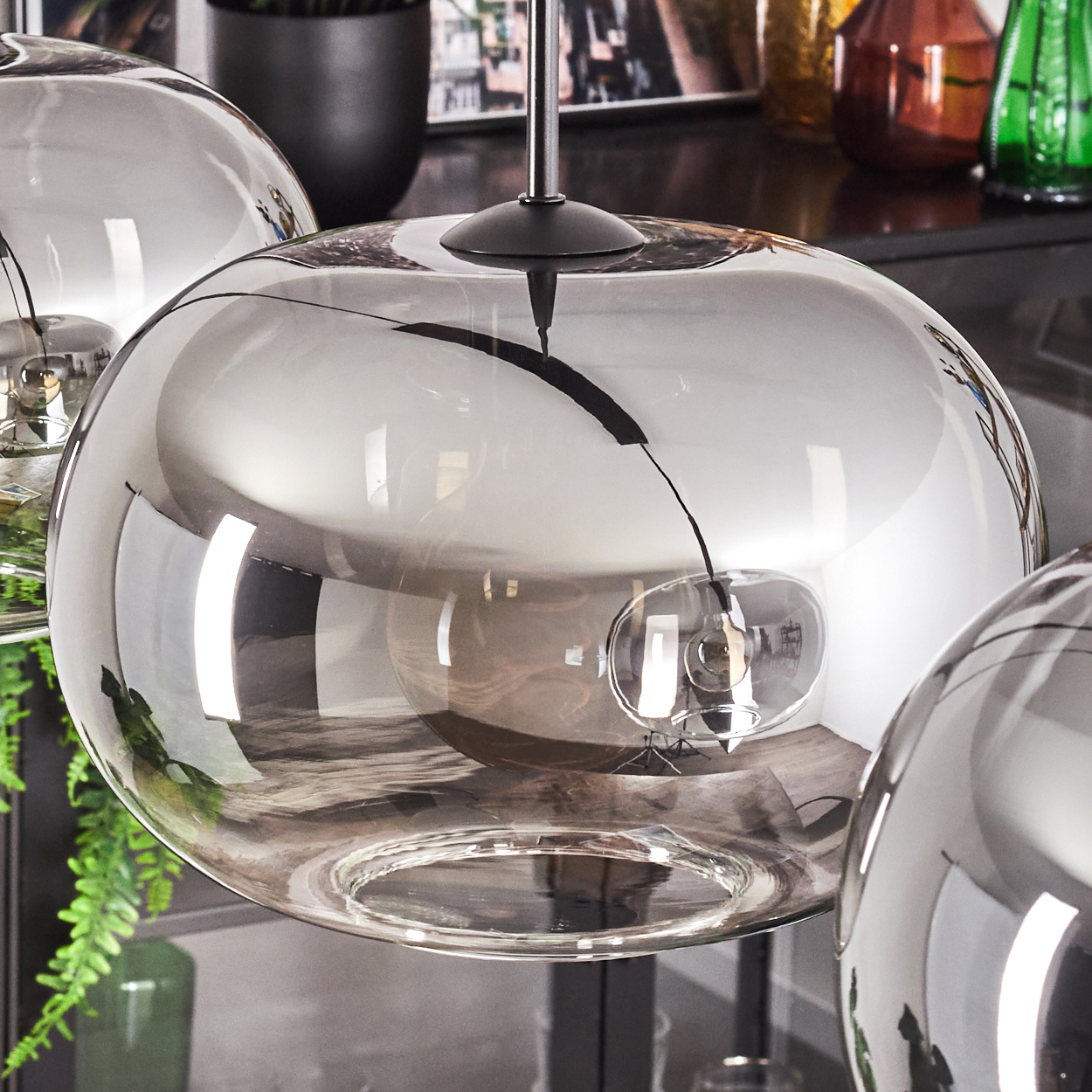 Nahaufnahme von drei Glaskugel-Pendelleuchten. Das Glas ist klar mit einer reflektierenden, rauchigen Beschichtung. Hintergrund hat Pflanzen und andere Dekorationen.