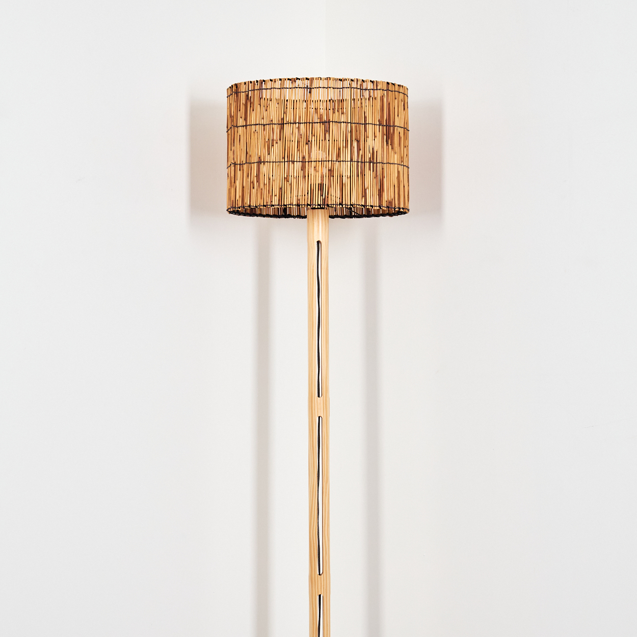 Eine Stehlampe mit zylindrischem Lampenschirm aus Bambusstäben. Der Ständer ist aus Holz, der Hintergrund ist eine weiße Wand.