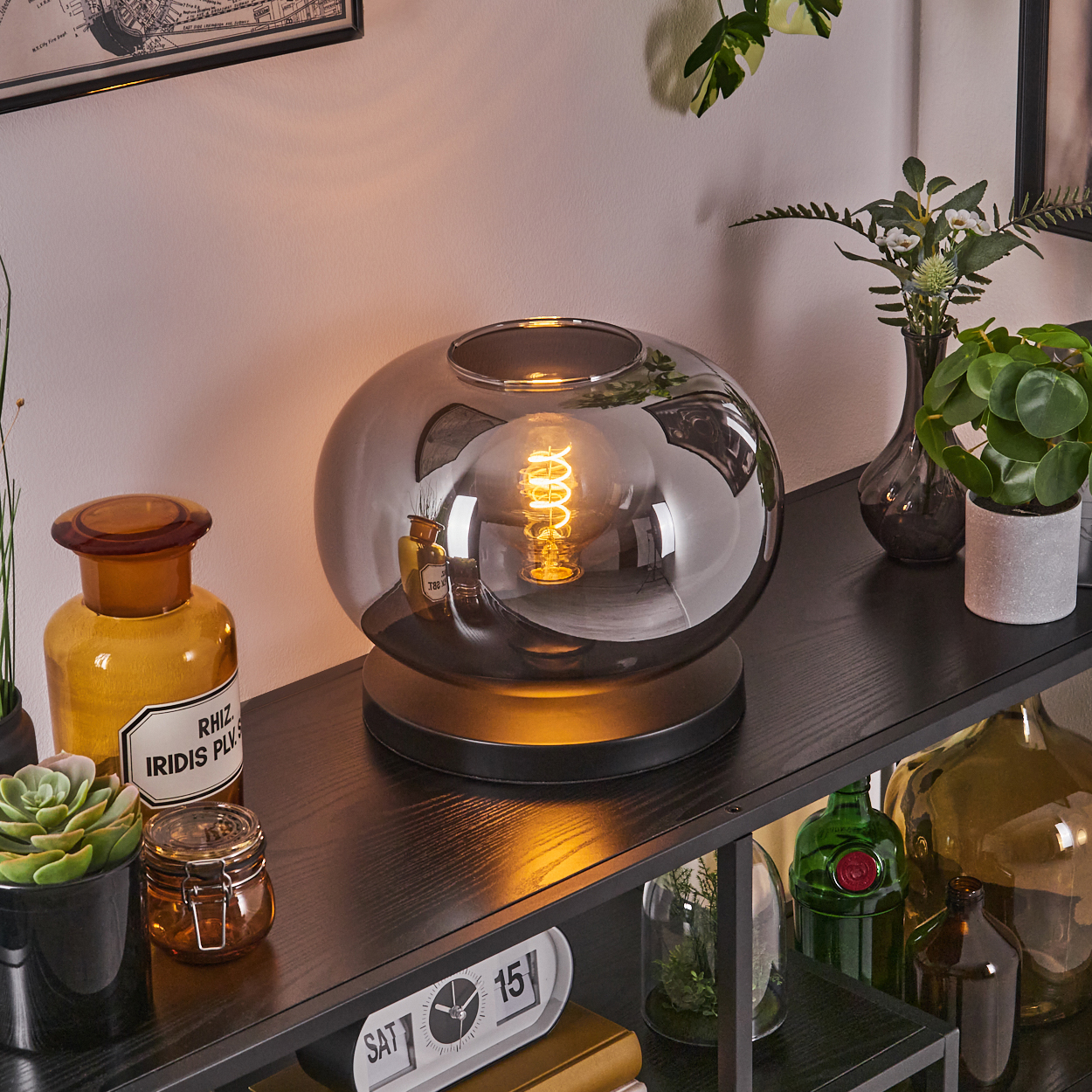 Ein dekoratives Regal mit einer beleuchteten Lampe, Gläsern, Pflanzen und einer Uhr. Die Lampe spiegelt die Umgebung wider.
