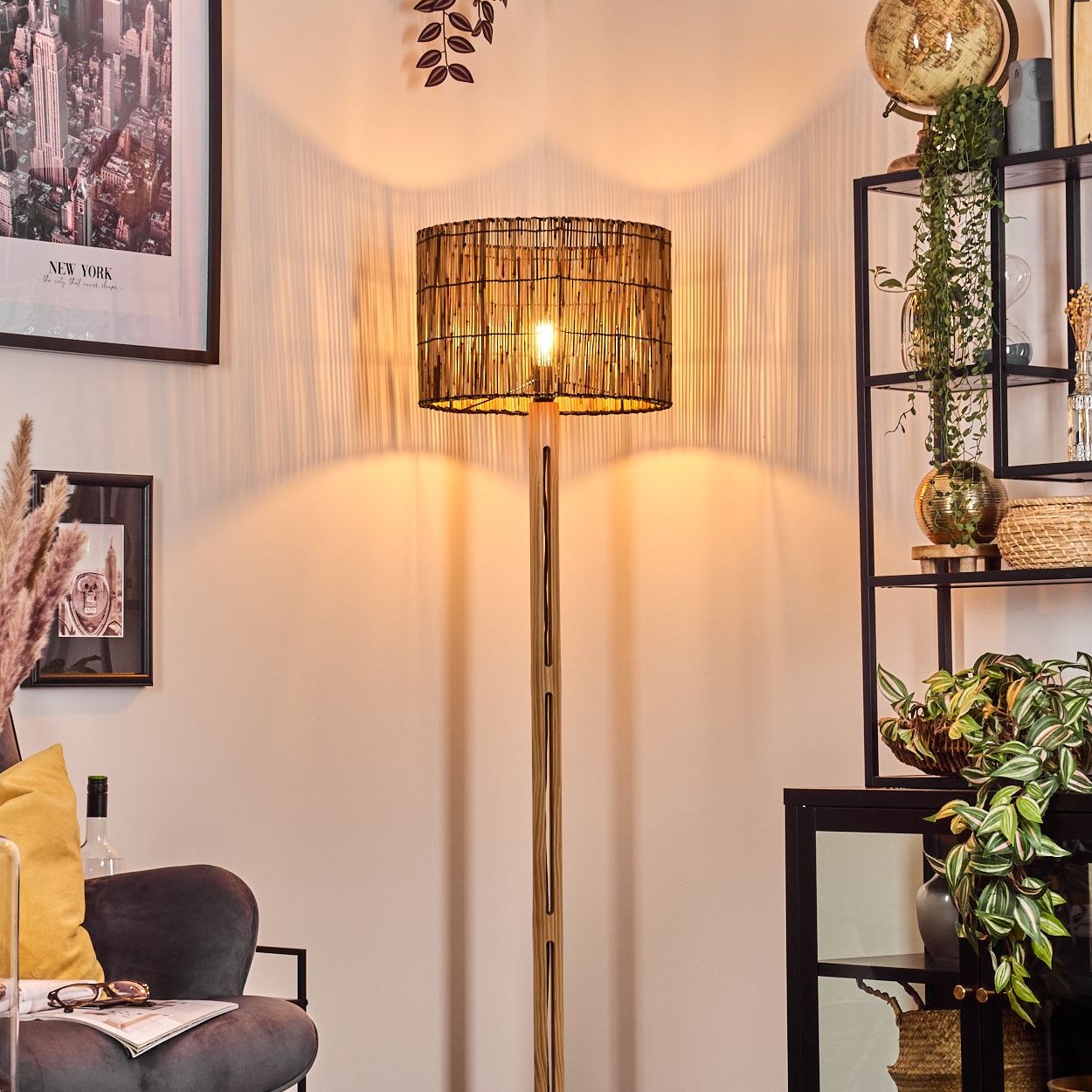 Eine beleuchtete Stehlampe mit gewebtem Schirm steht in einem Raum. Ein schwarzes Regal mit Dekoration und ein Stuhl mit gelbem Kissen sind in der Nähe.