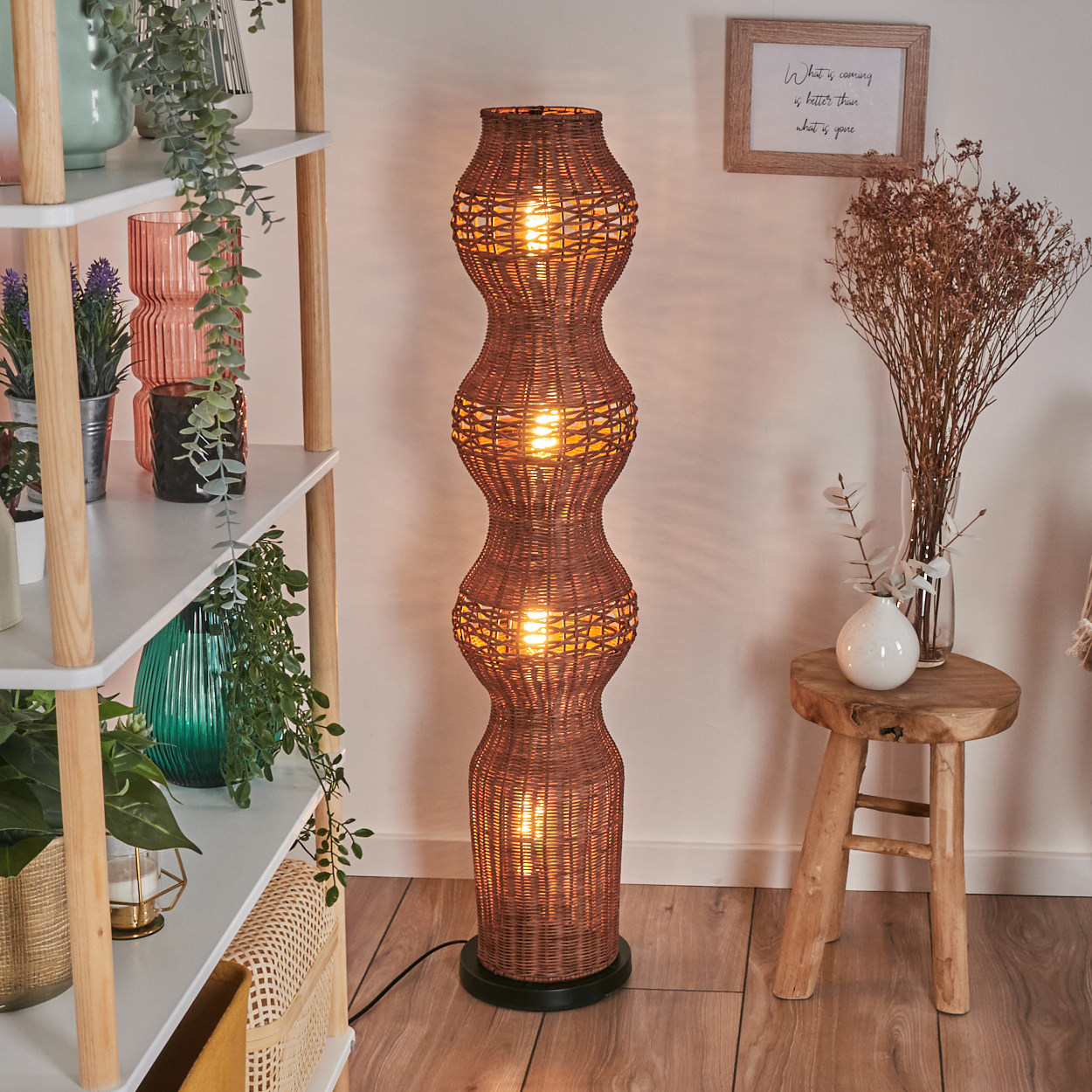 Eine gewebte Stehlampe, ein Holzhocker mit Vase und ein gerahmtes Zitat. Regale mit Pflanzen im Hintergrund.