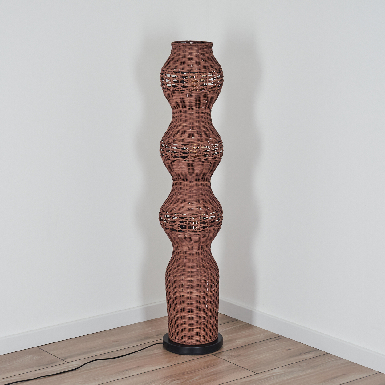Eine hohe, braune Korblampe steht in einer Ecke vor weißen Wänden. Sie hat einen schwarzen Sockel und einen Holzboden.