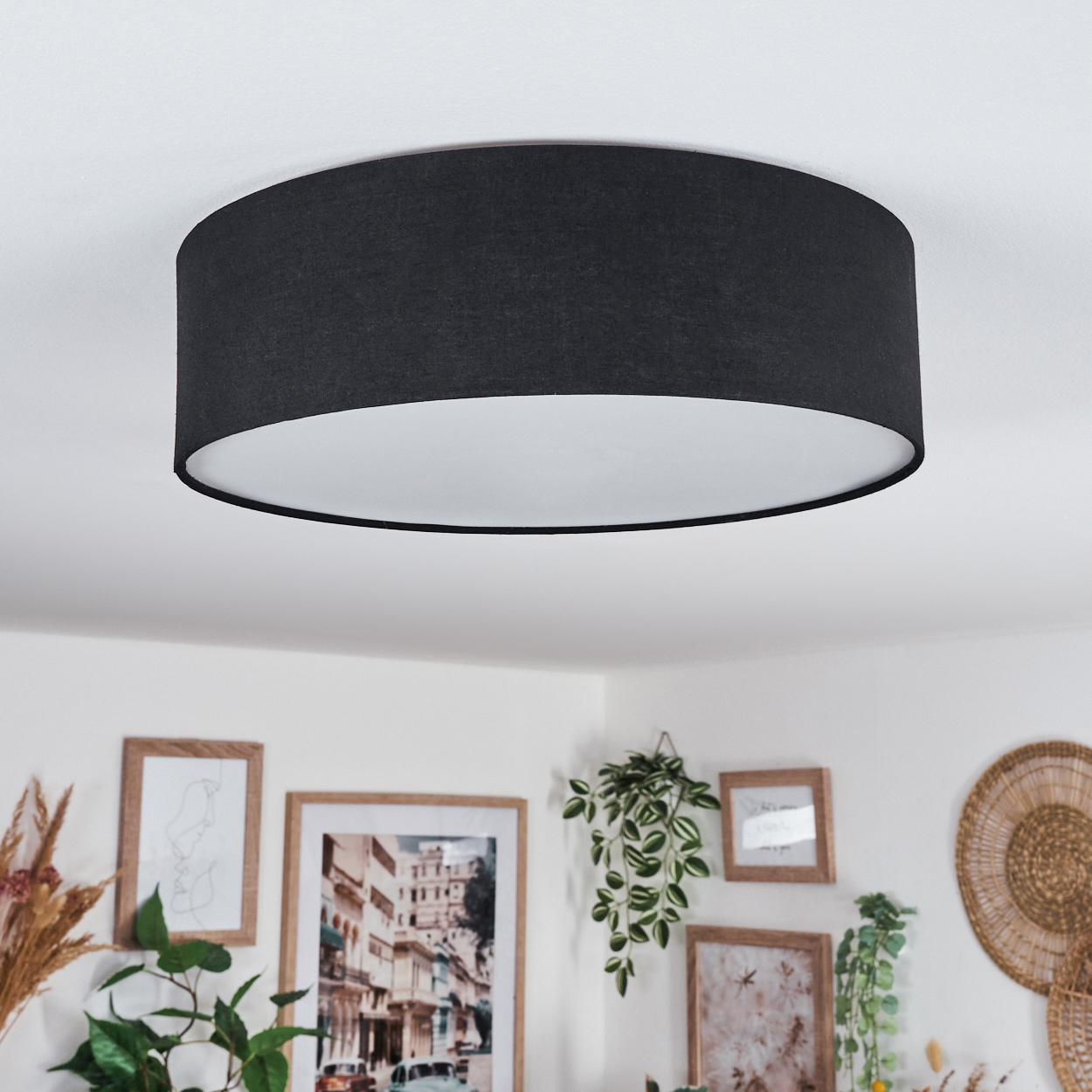 Eine schwarze Deckenlampe mit weißem Innenraum. Darunter schmücken gerahmte Bilder und Pflanzen die weiße Wand.