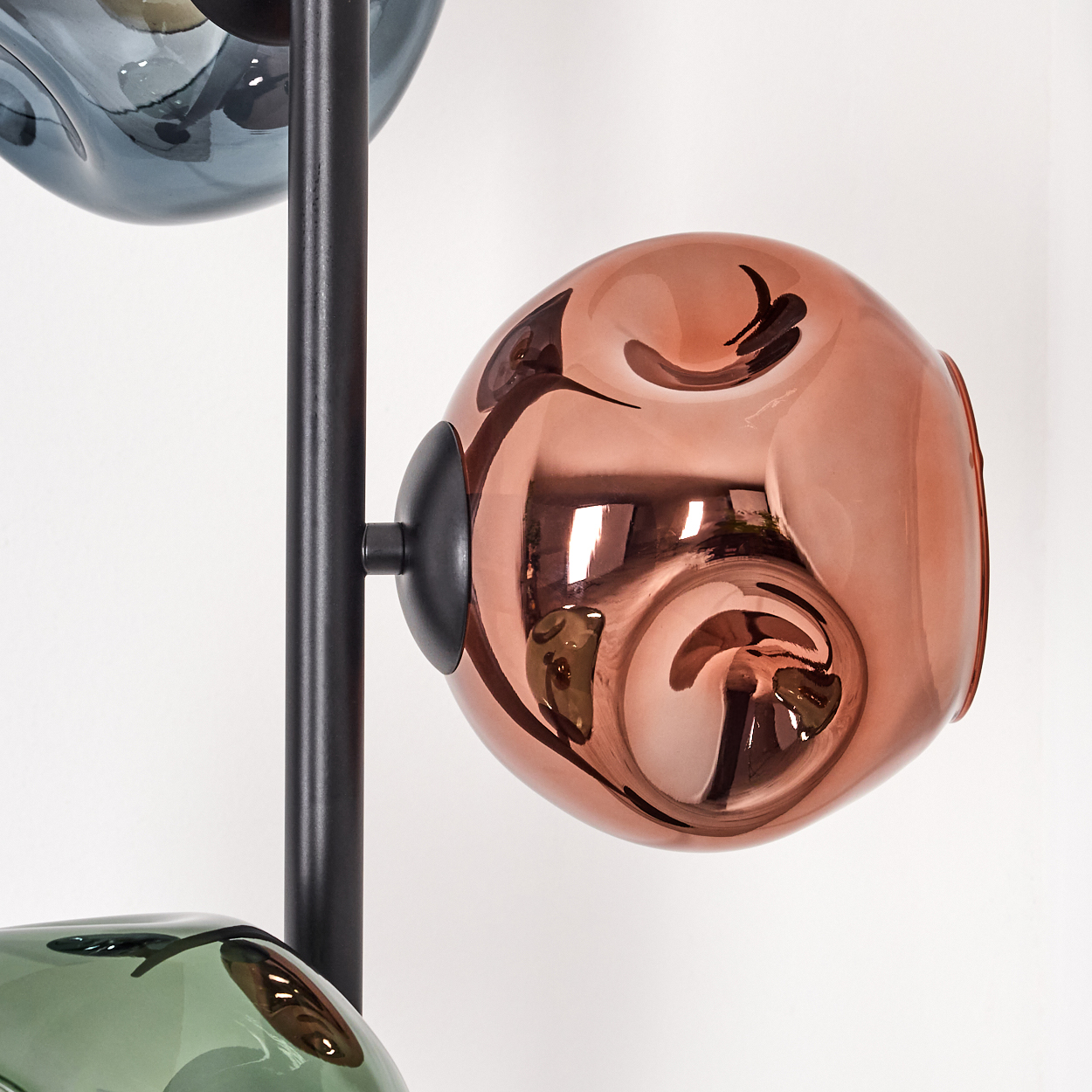 Eine moderne Lampe mit drei Glaskugeln in Blau, Kupfer und Grün, die an einer schwarzen Stange befestigt sind, vor weißem Hintergrund.
