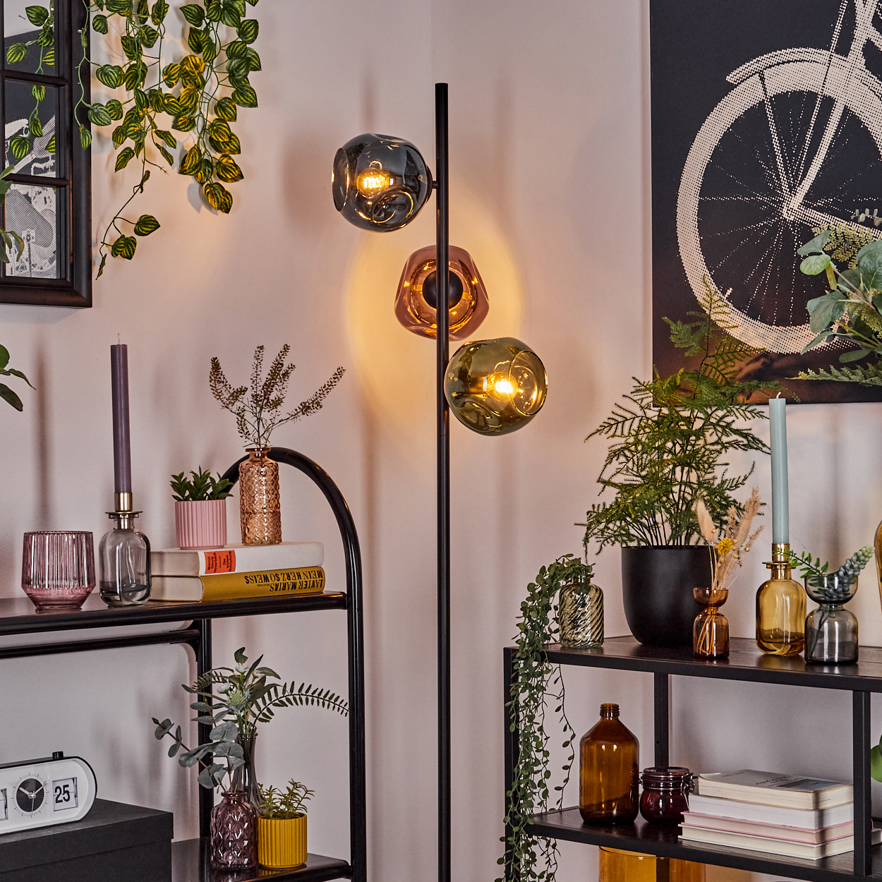 Eine Zimmerecke mit einer schwarzen Stehlampe mit drei Glaskugeln, Regalen mit Pflanzen und Dekoration sowie einem Fahrradkunstwerk.