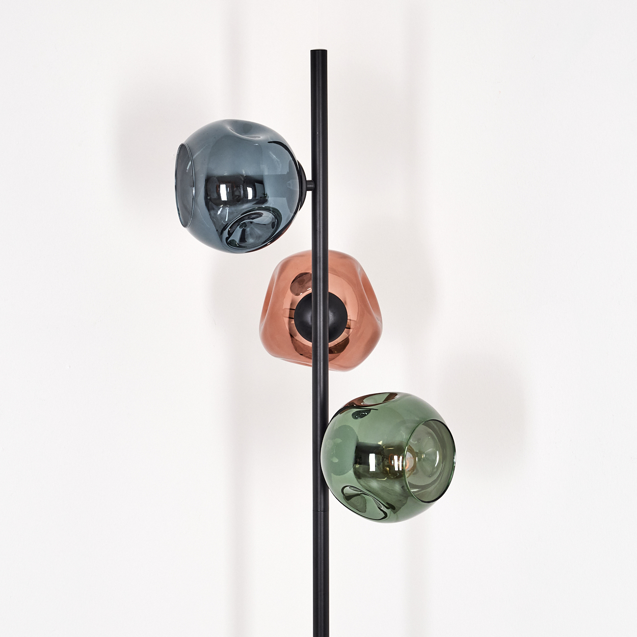 Eine moderne Lampe mit drei farbigen Glasschirmen: blau, kupfer und grün. Sie hat eine schwarze Stange und steht vor einer weißen Wand.