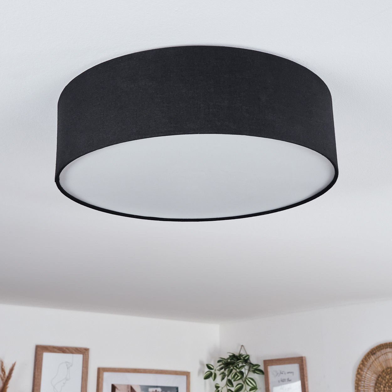 Eine schwarze kreisförmige Deckenlampe mit weißem Innenraum. Der Hintergrund zeigt gerahmte Bilder, Pflanzen und eine Wanddekoration.