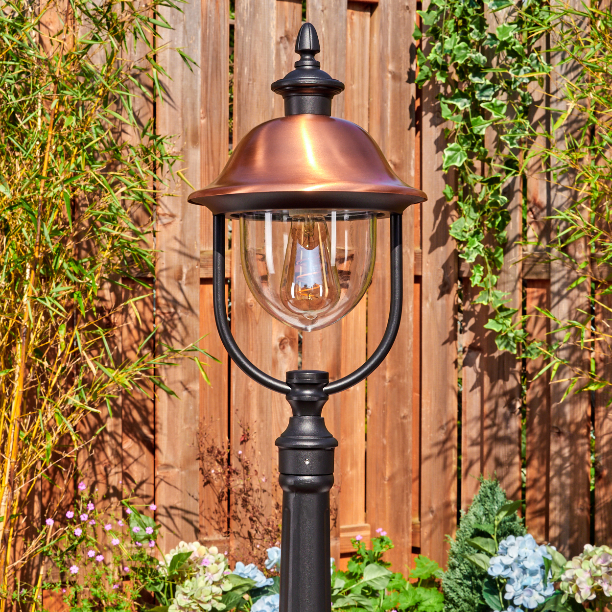 Eine dekorative Außenlampe mit kupferfarbener Oberseite und Glaskugel. Sie steht vor einem Holzzaun und ist von Pflanzen und Blumen umgeben.